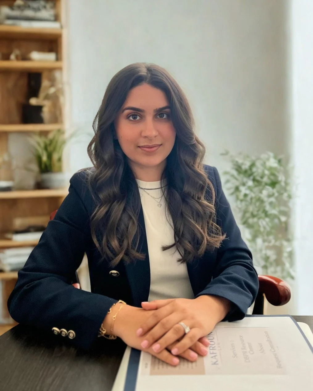 A woman with long curly brown hair sitting at a table in an office. She is wearing a black blazer and a white shirt, with her hands clasped on the table. There is a document in front of her and bookshelves and plants in the background.