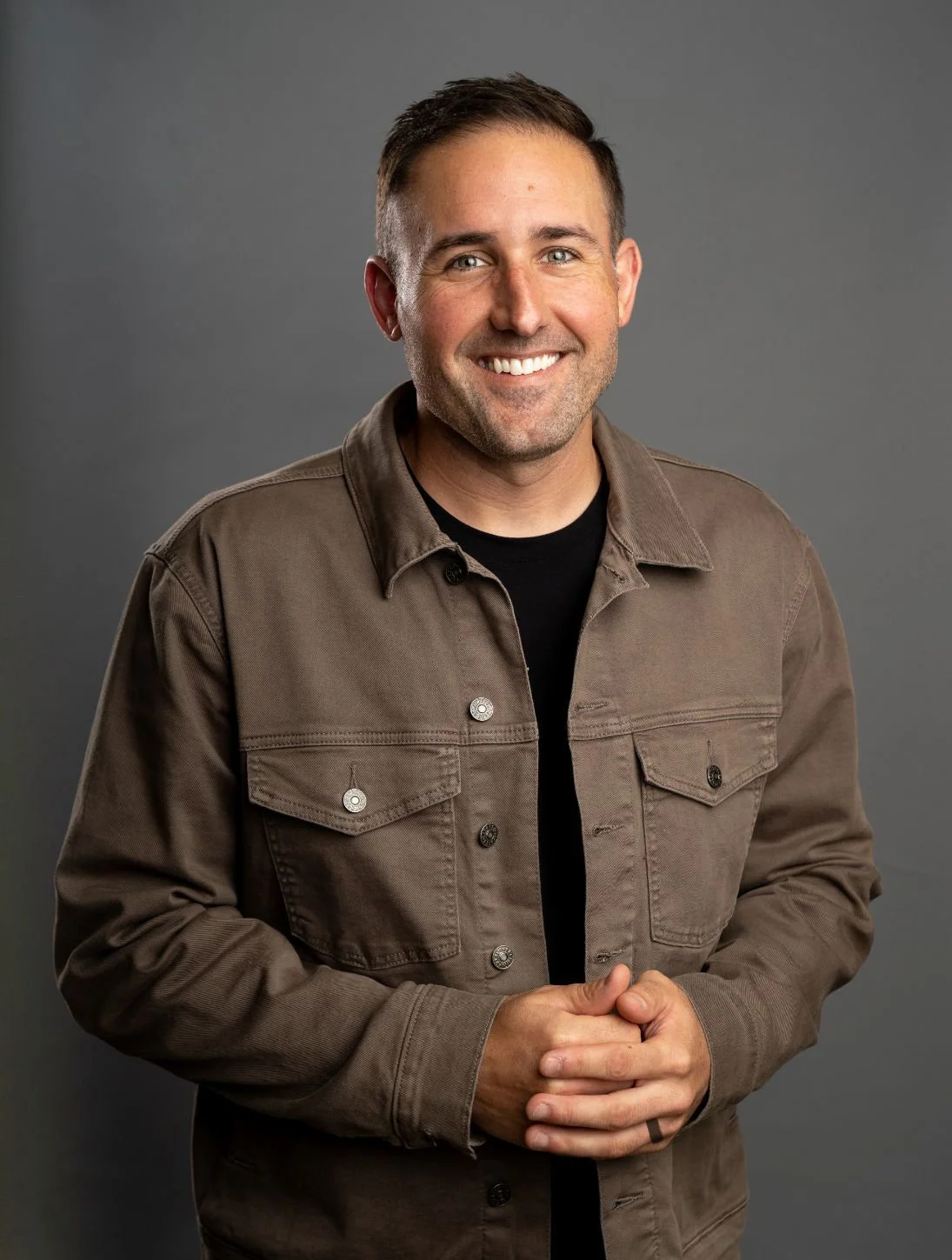 A man with short brown hair, blue eyes, and a beard, smiling, wearing a brown jacket over a black shirt, standing against a gray background.