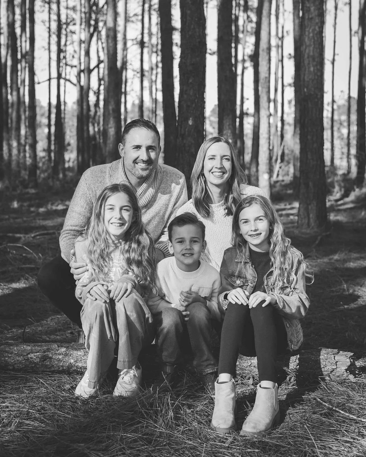 A black and white photograph of a family of six in a forest, consisting of two adults and four children, smiling at the camera.