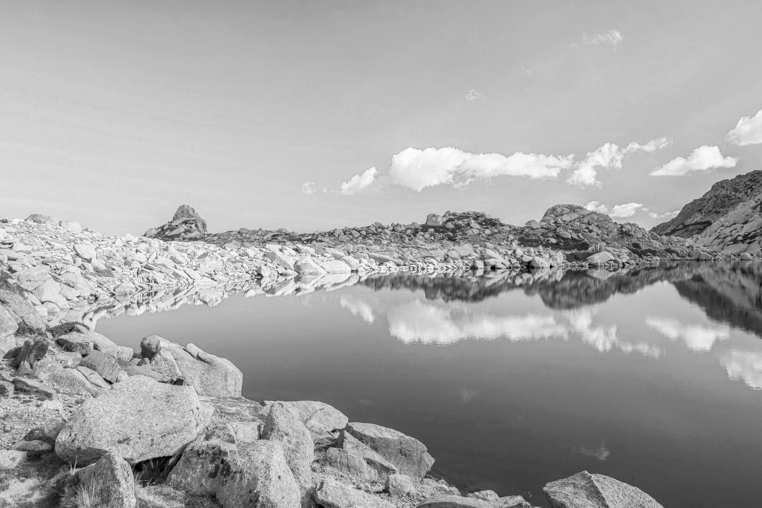 Black and white photograph of a rocky landscape with large stones along the shoreline and a calm body of water, reflecting the rocks and sky above, with distant mountains and a partly cloudy sky.