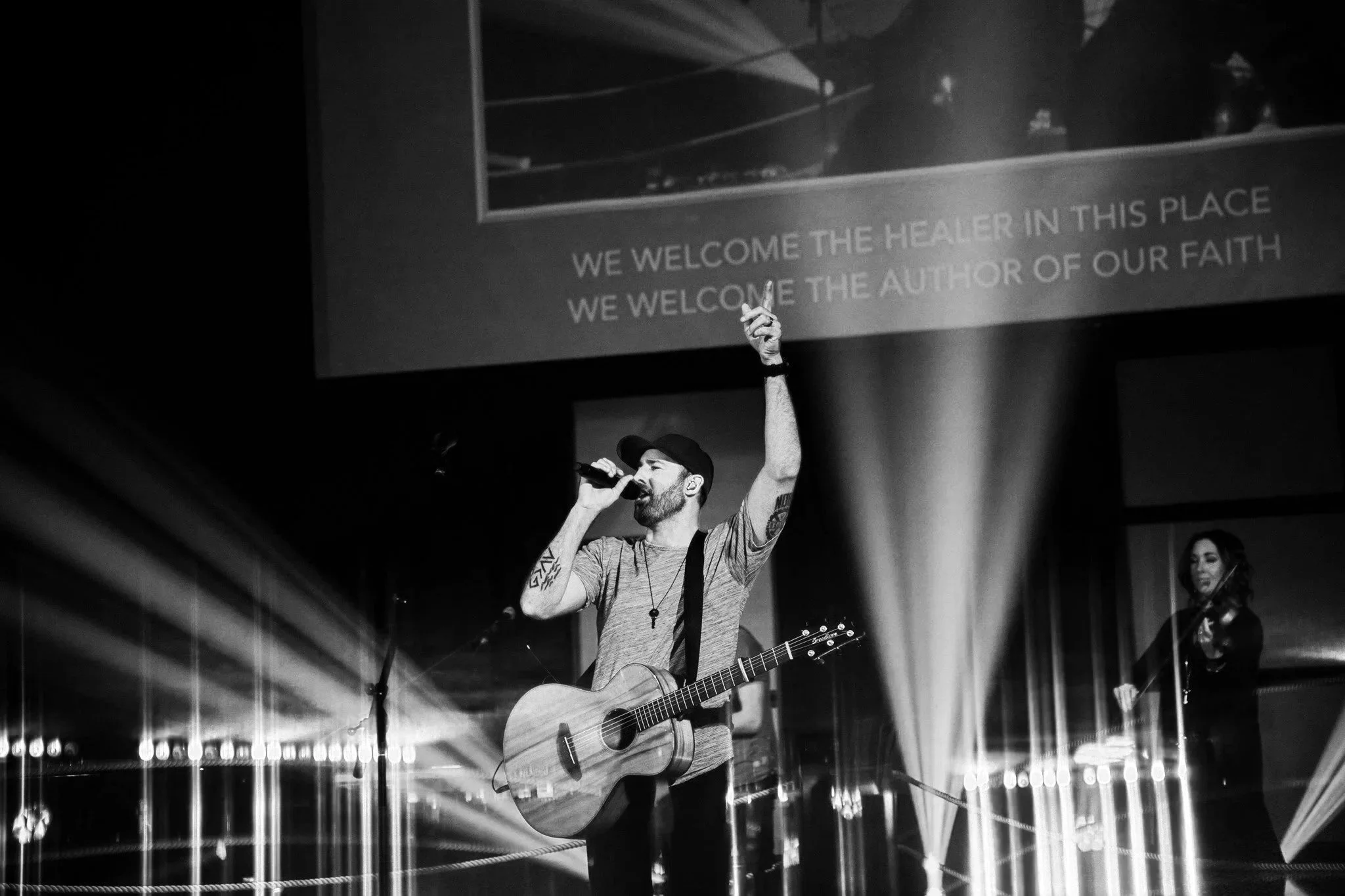 A man with a beard, wearing a cap, holding a microphone and a guitar, singing on stage with a woman playing a violin in the background, illuminated by stage lighting, with a projected screen that reads "We welcome the healer in this place, we welcome the author of our faith."