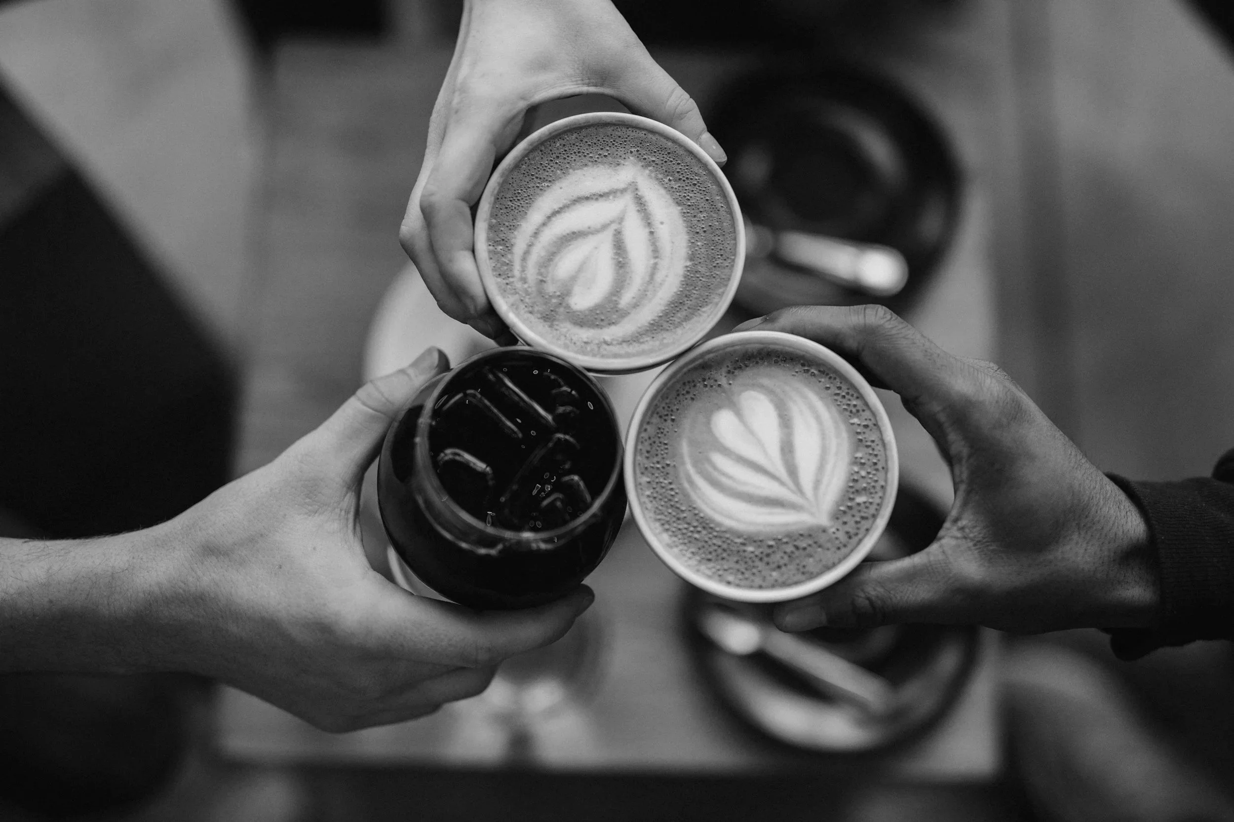 Three people clinking cups of coffee with latte art on top, one hand holding a glass of iced coffee, in black and white.