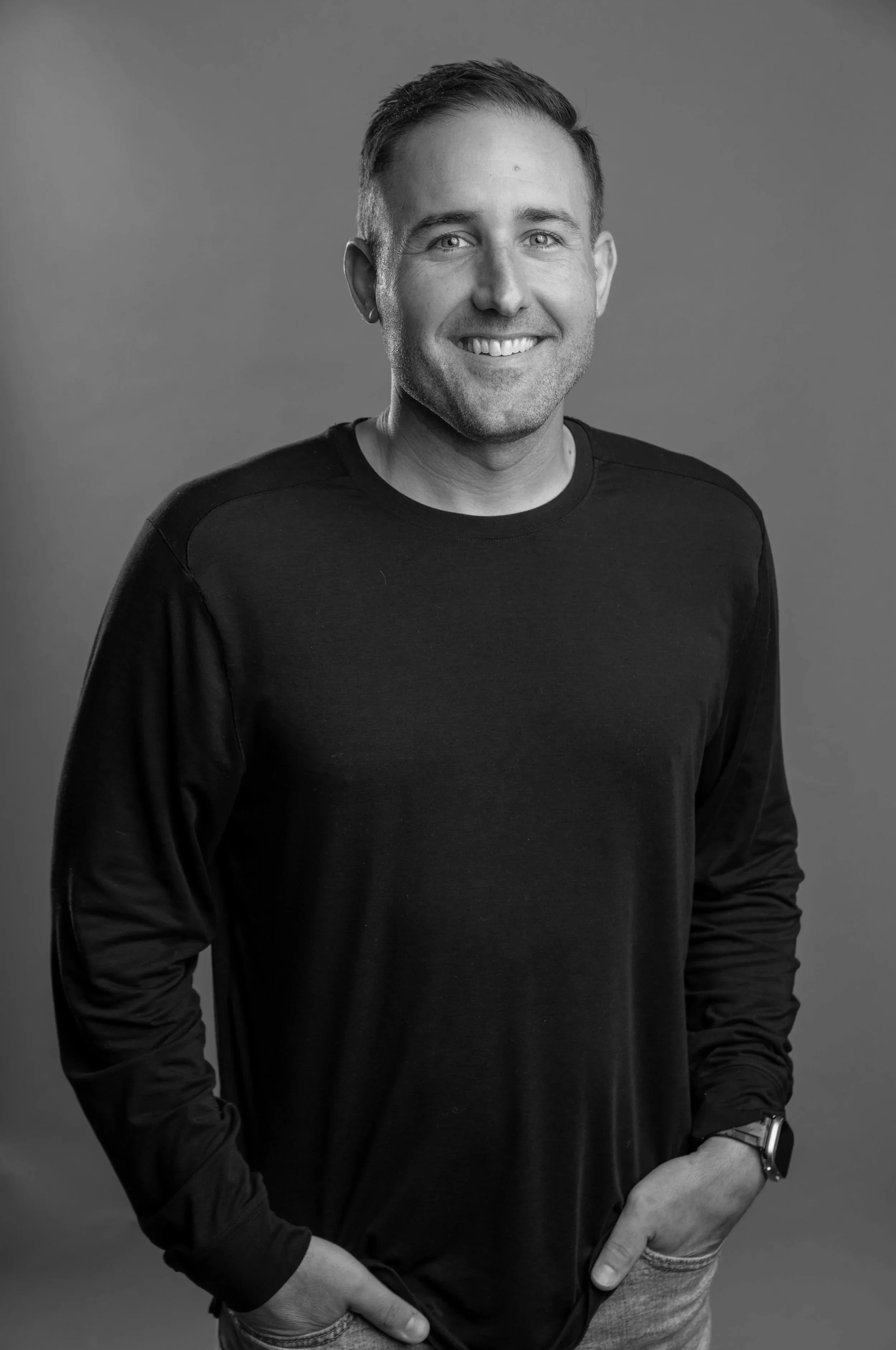 A smiling man wearing a black long-sleeve shirt and a watch, standing with his hands in his pockets, in a studio setting with a plain background.