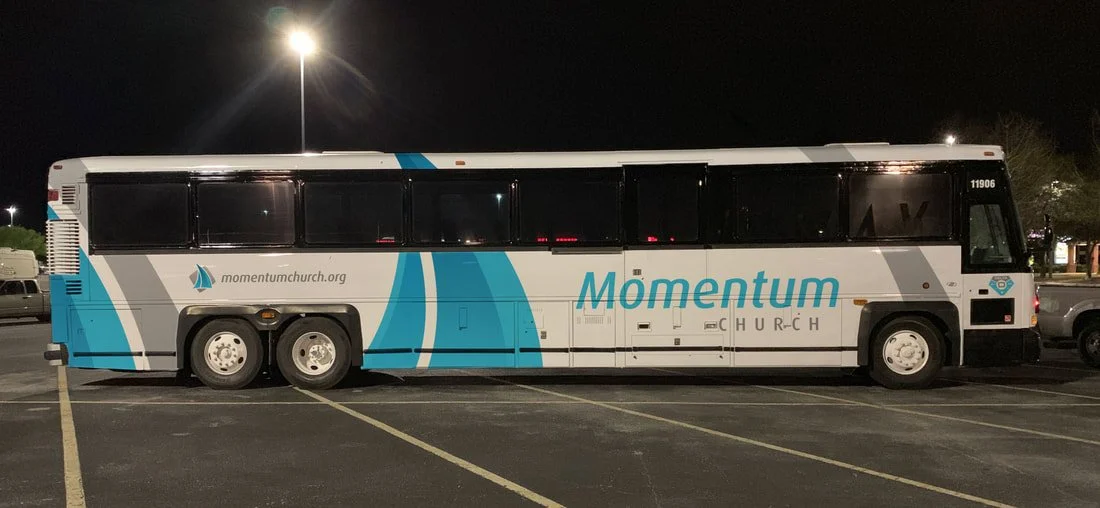 A bus with a white and blue exterior parked in a lot at night, with the words 'Momentum' and 'momentumchurch.org' on its side.