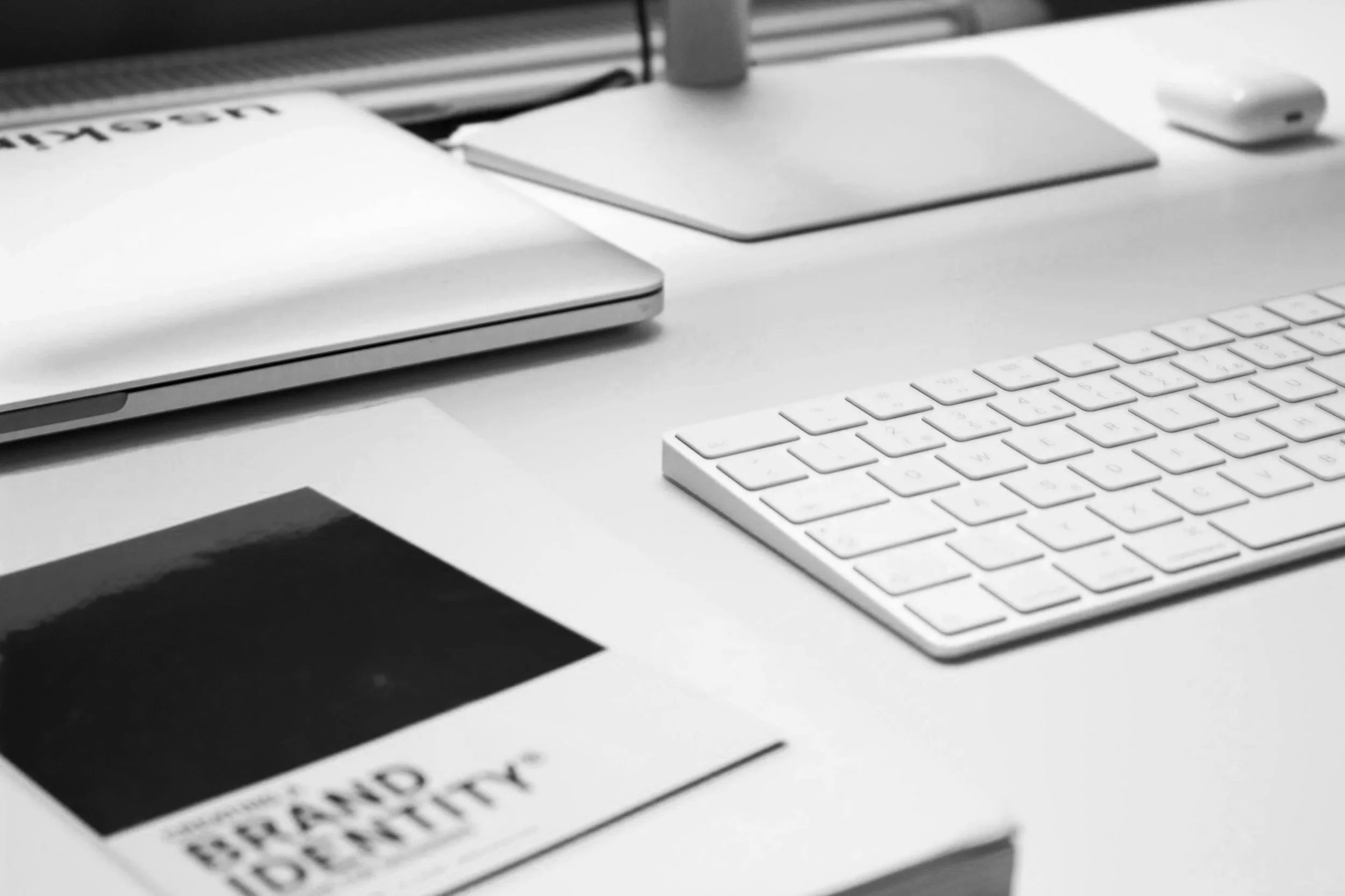 A white computer keyboard, a closed laptop, a magazine, a white mouse, and a notepad on a white desk in an office setting.