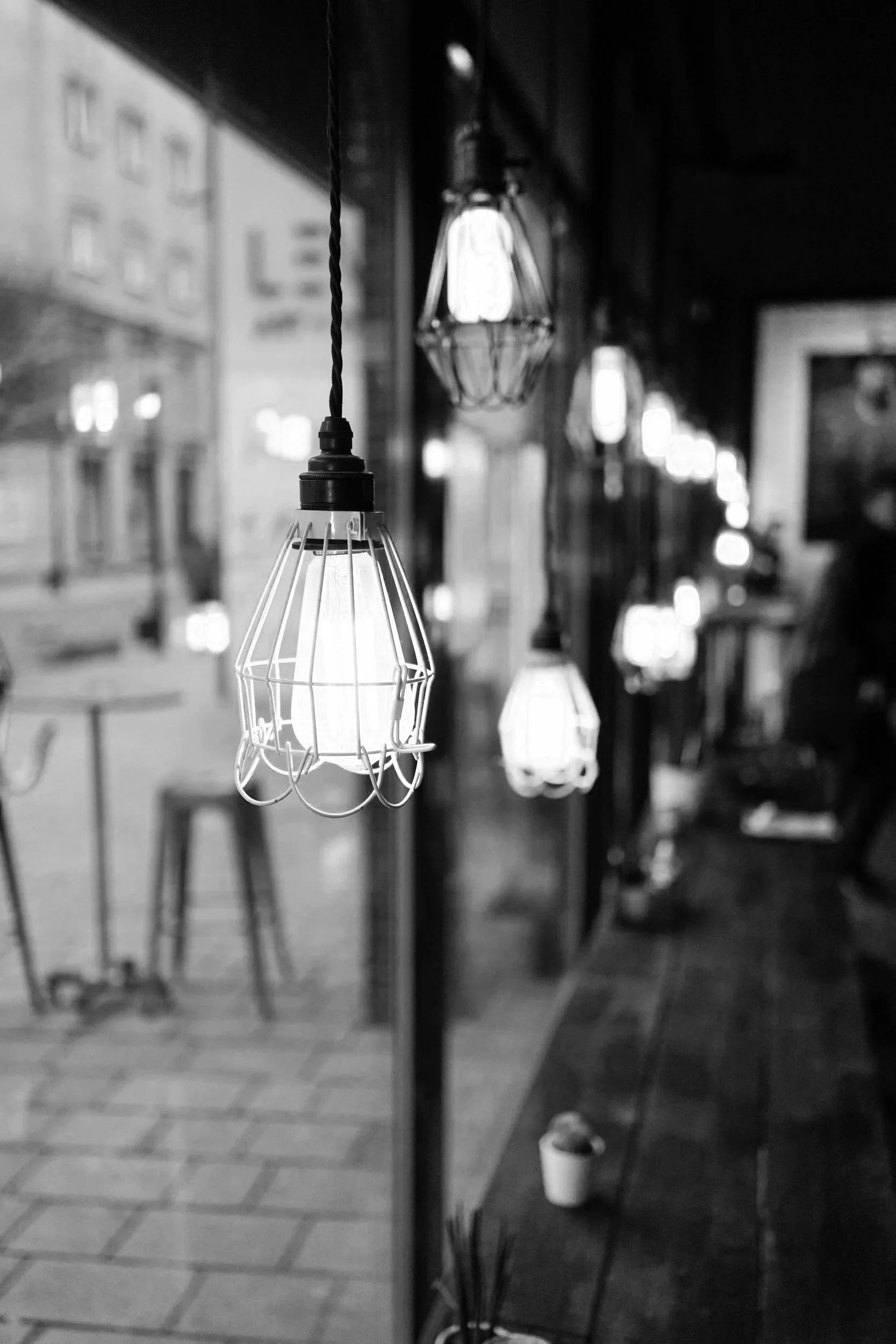 A row of hanging pendant lights with wire cages inside a dimly lit room or cafe, some with visible filaments, with a blurred background including tables and chairs.