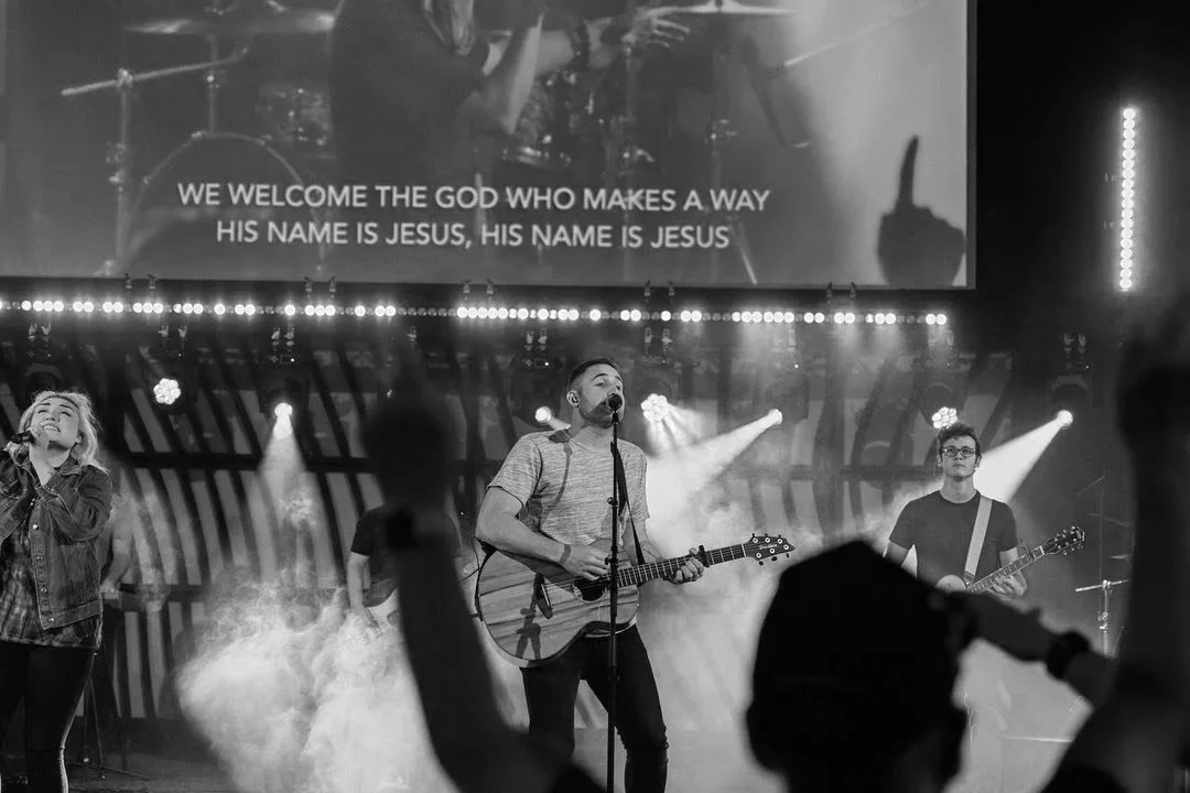A black and white photo of a live musical performance with three musicians. A man in the center is playing an acoustic guitar and singing into a microphone. A woman on the left is singing, and another man on the right is playing an electric guitar. There is a large screen above them with the lyrics: "We welcome the God who makes a way. His name is Jesus, his name is Jesus." Audience members can be seen in the foreground raising their hands.