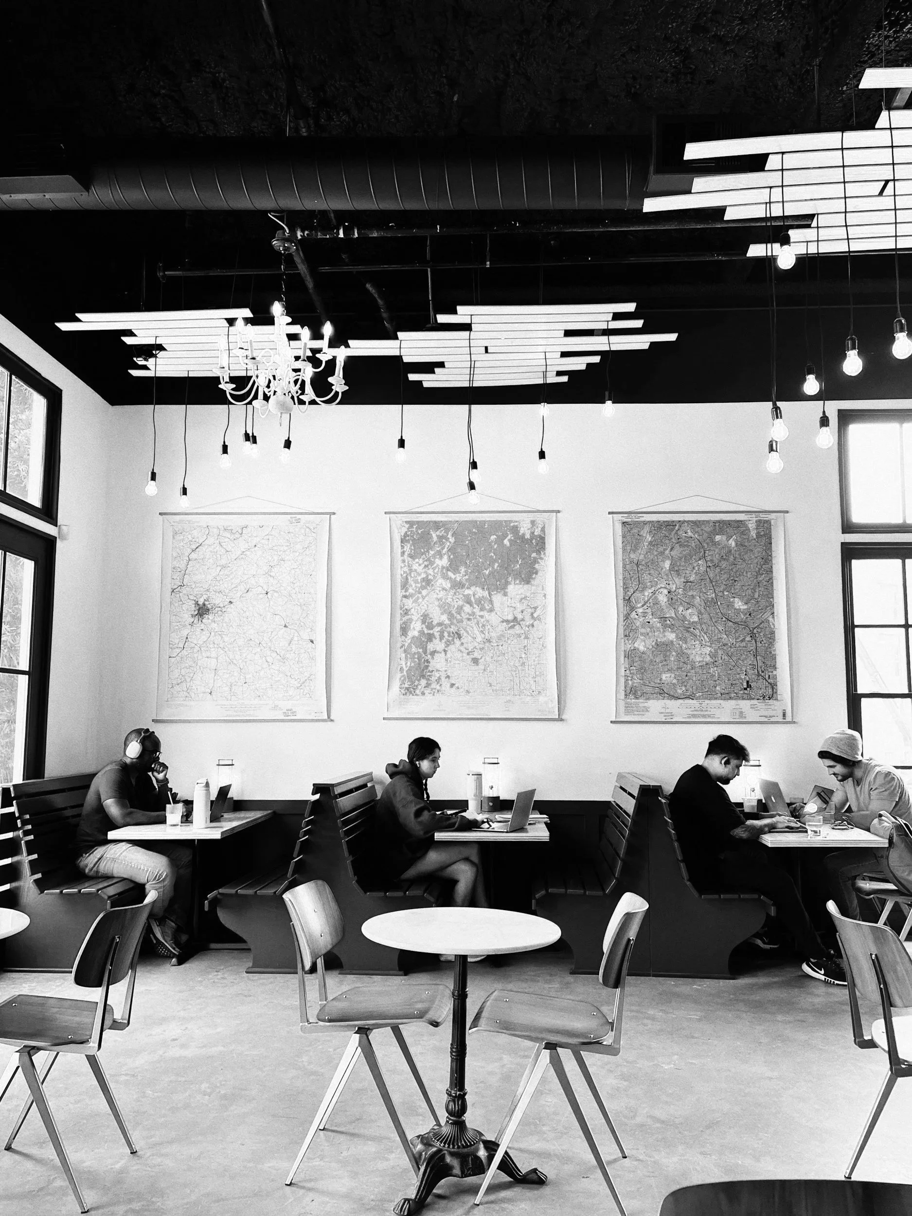 Black and white photo of a cafe interior with four people seated at a long table working on laptops, two women and two men, with maps on the wall behind them, large windows on the sides, and unique light fixtures on the ceiling.