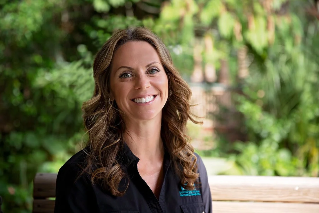 A woman smiling outdoors, seated on a park bench with greenery in the background.