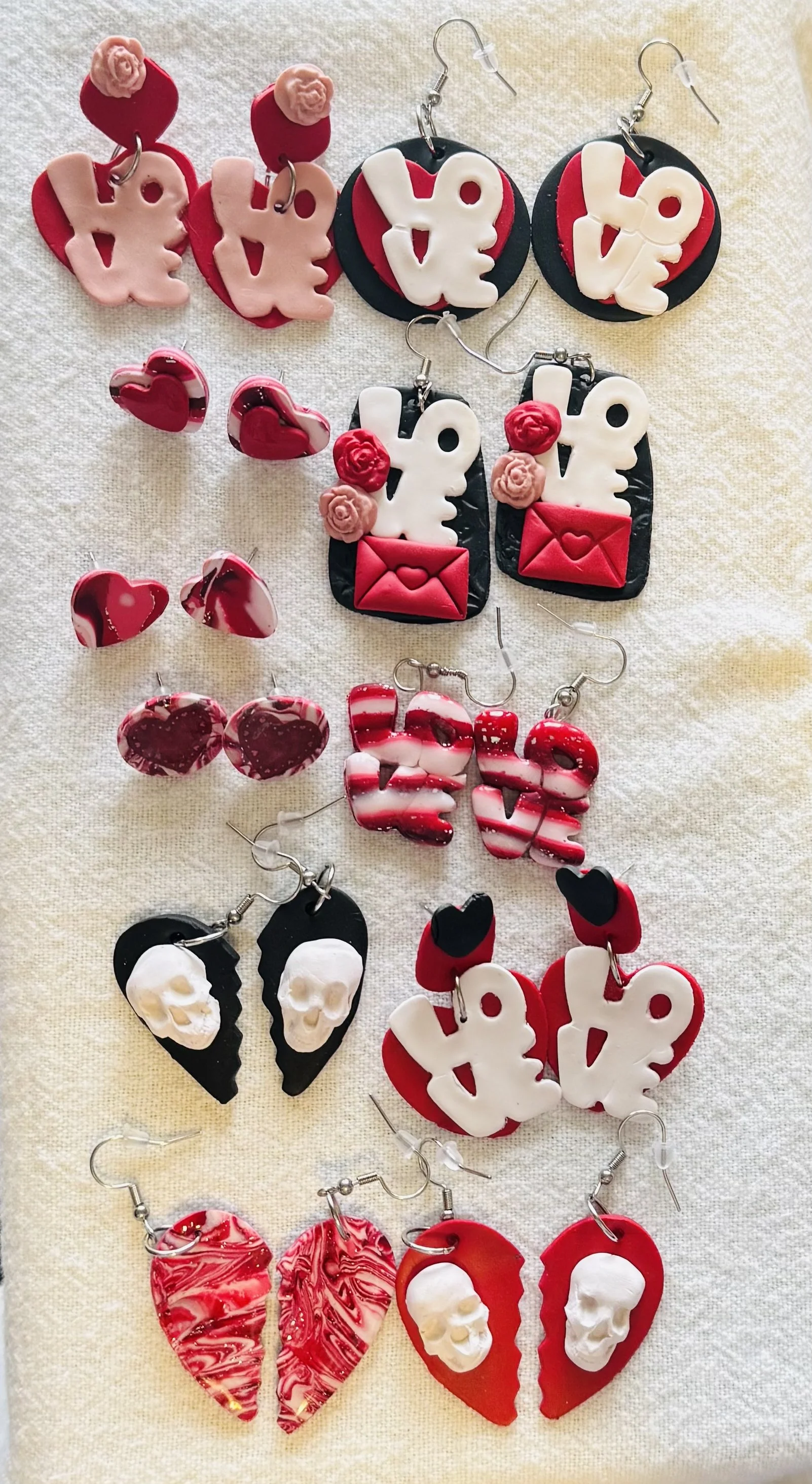 Assorted earrings in Valentine's Day theme featuring hearts, the word "love," skulls, and marbled designs, arranged on a light fabric background.