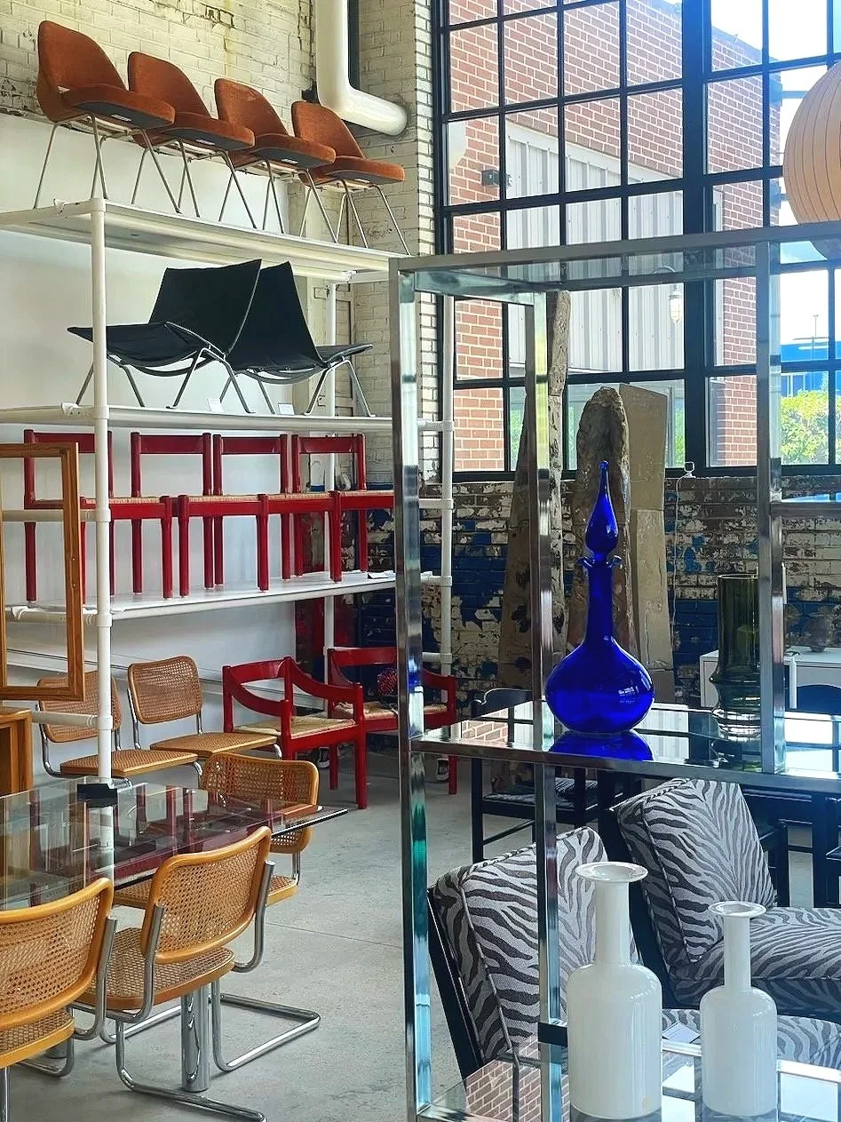 Interior of a modern furniture showroom with shelves displaying colorful chairs, a table with zebra-patterned upholstered chairs, and decorative glass vases, near large windows allowing natural light.