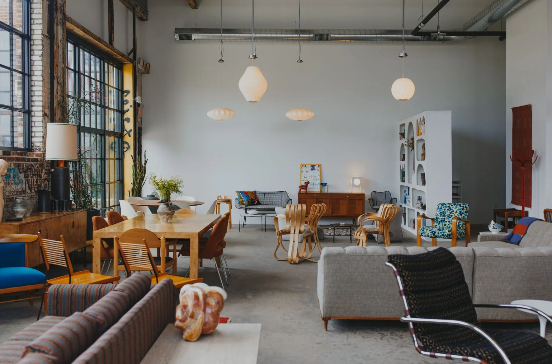 Interior of a stylish, open-concept living room and dining area with modern decor. Features a large window with black frames, various chairs including a gray sofa, a wooden dining table with chairs, wall shelves, and hanging pendant lights.