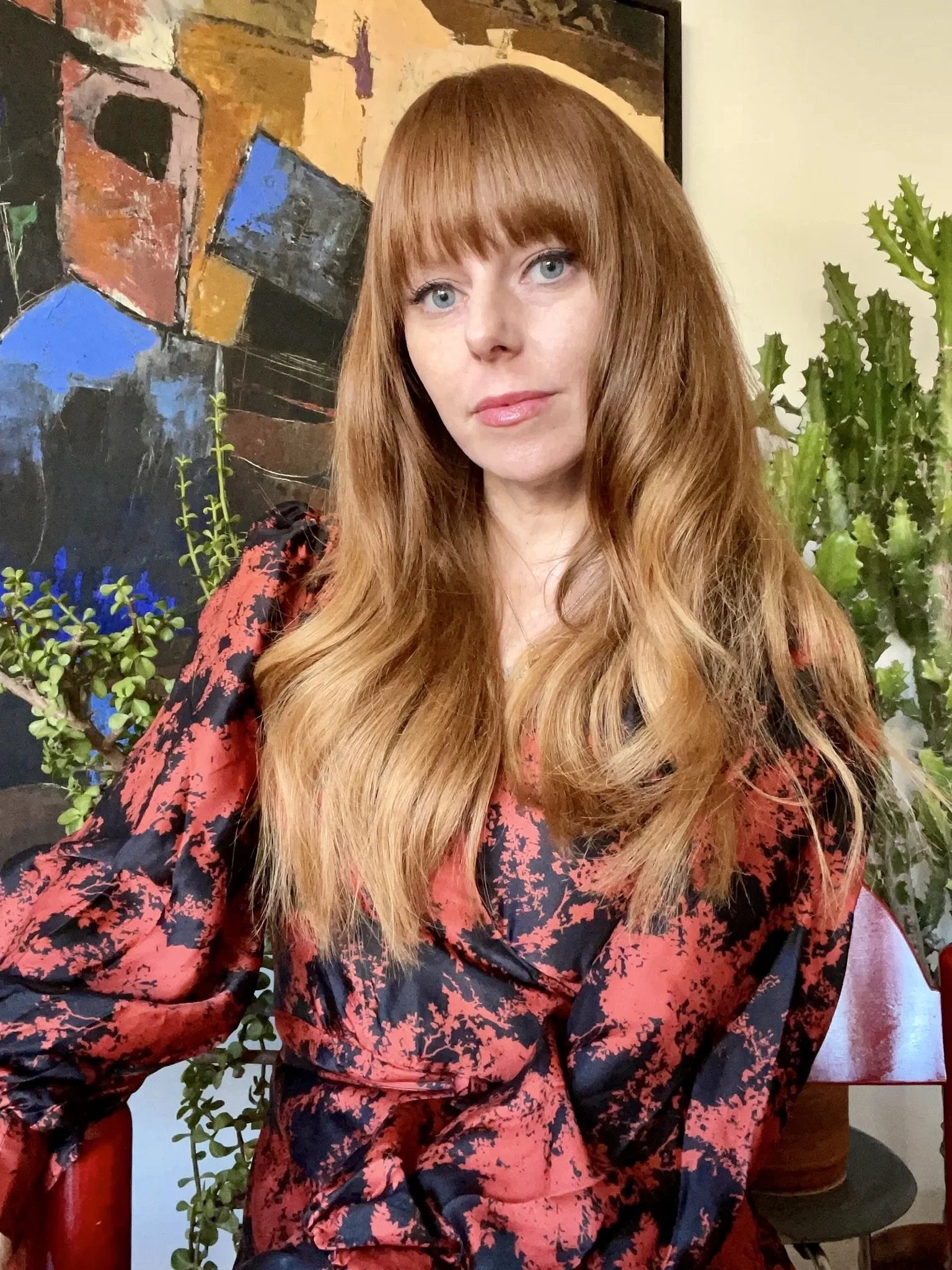 A woman with long red hair and bangs, wearing a red and black patterned blouse, sitting in front of abstract artwork and green plants.