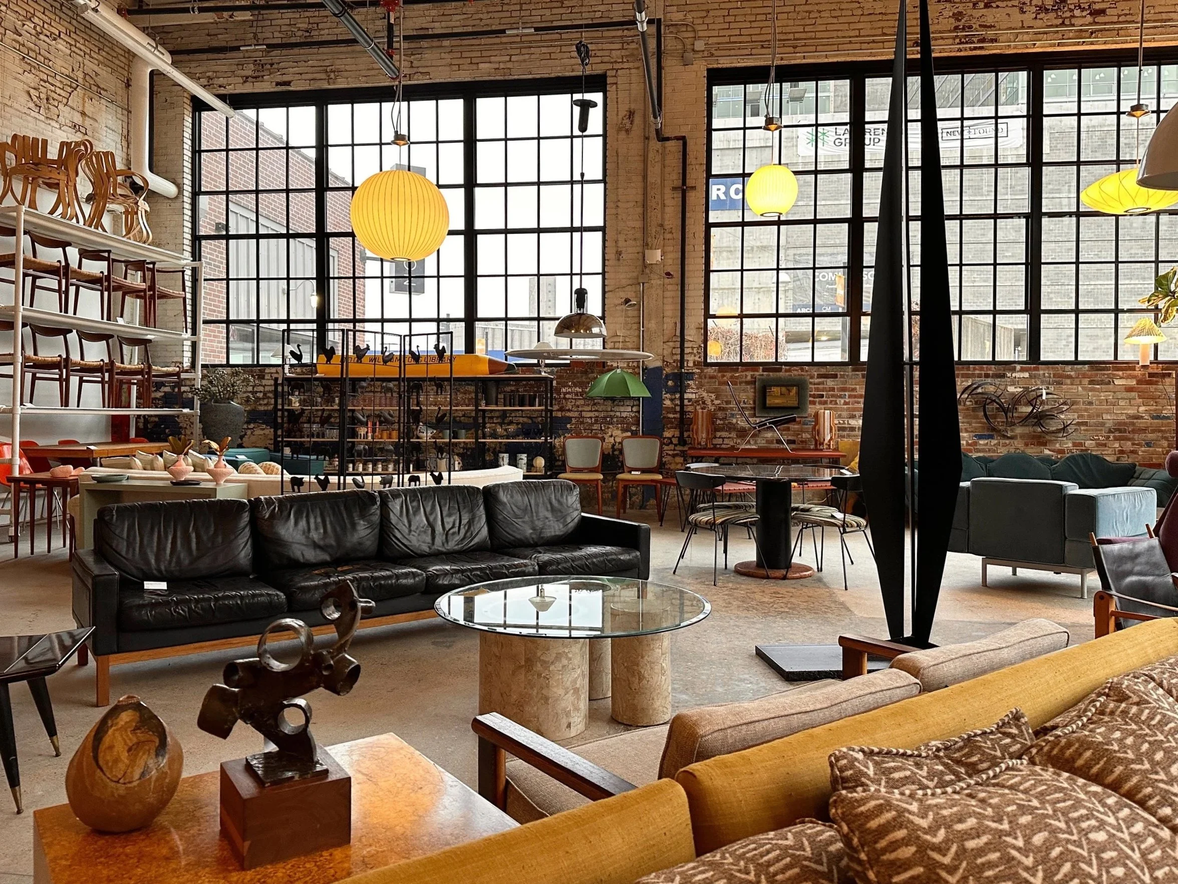 Interior view of a contemporary furniture store with large windows, exposed brick walls, and various chairs, sofas, tables, and decorative items on display, illuminated by pendant lights.