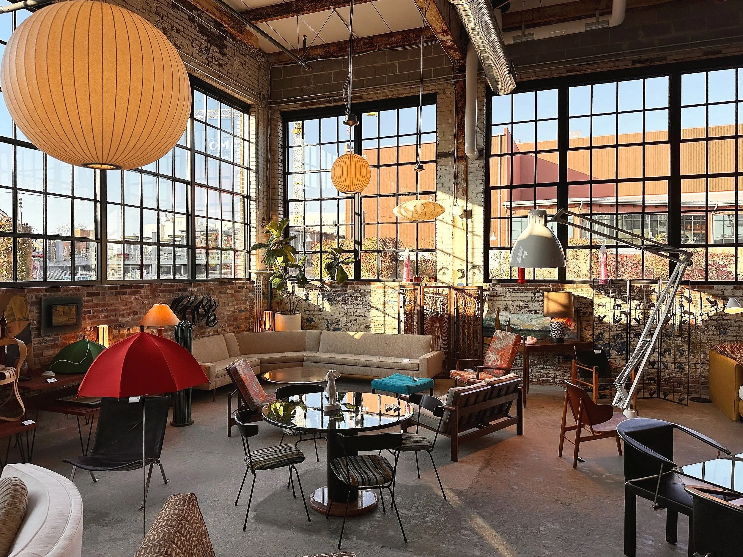 Interior view of a spacious, industrial-style living room with large windows, exposed brick walls, and eclectic furniture including a beige sectional sofa, black chairs, and various lamps and decor, with sunlight streaming in.