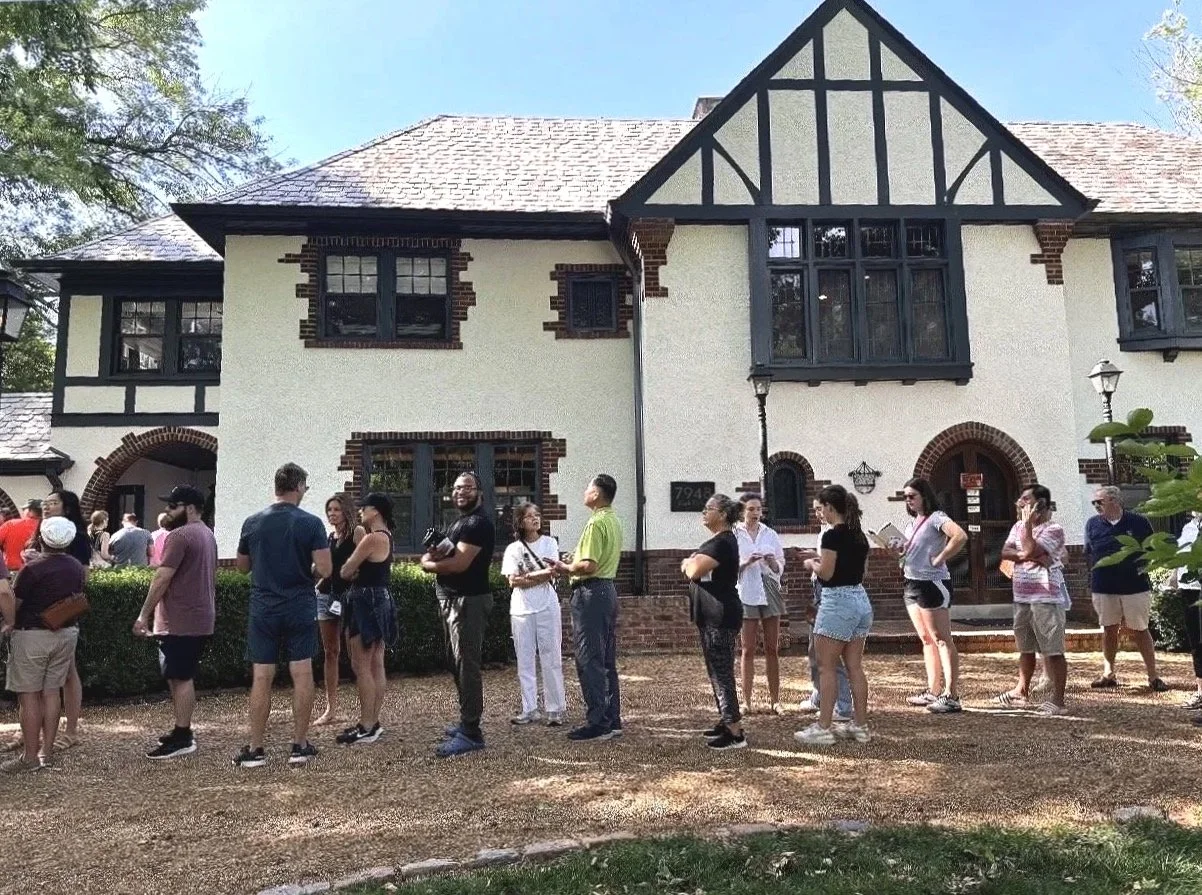 A group of people standing in line outside a large, Tudor-style house with a white facade, black trim, and brick accents.