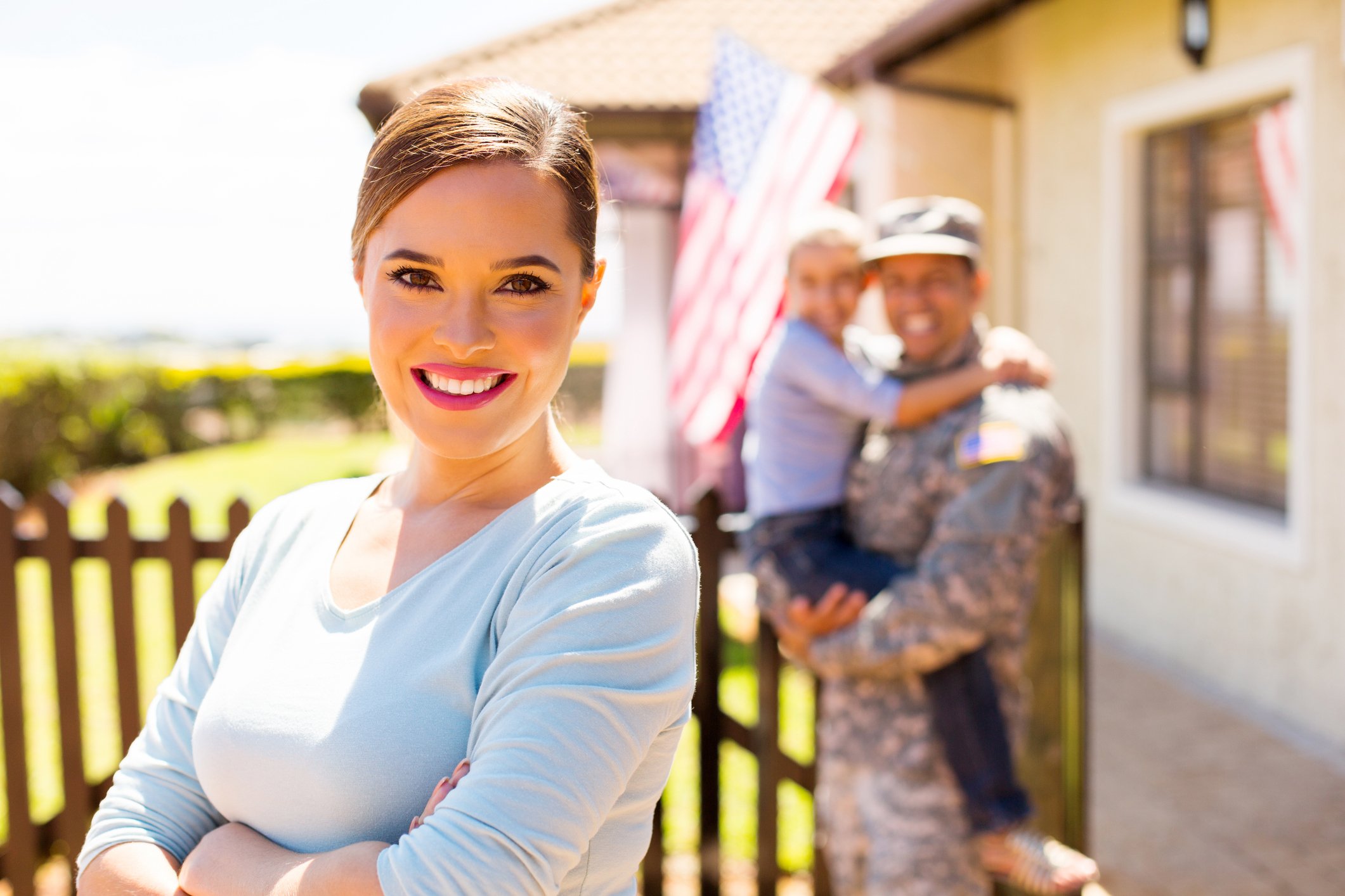 A military spouse smiling in the foreground with a man in military uniform and a young girl hugging him in the background outside a house decorated with American flags.