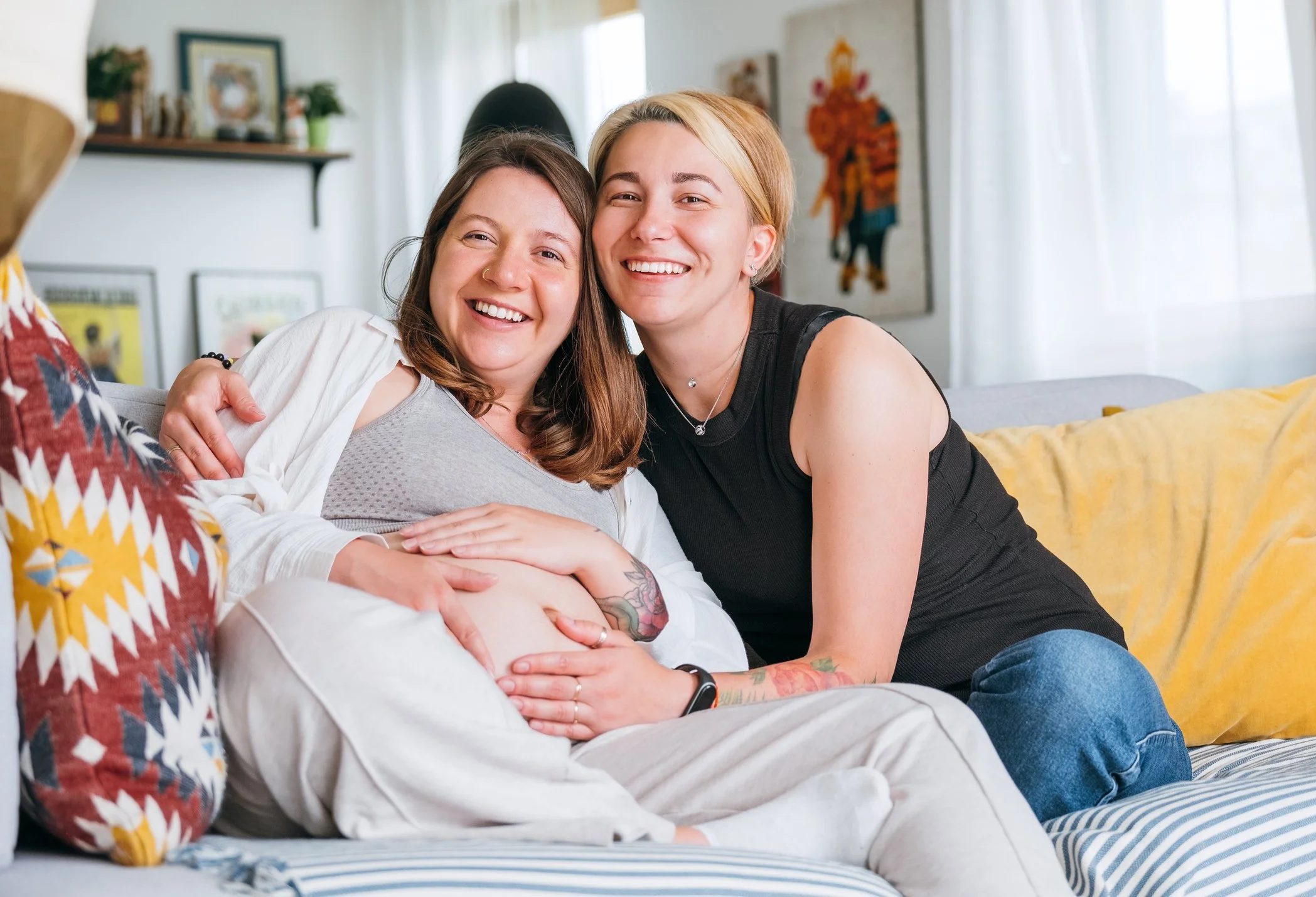 Two women sitting on a couch, smiling and hugging, in a cozy living room.