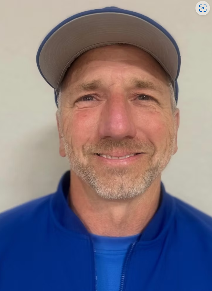 A middle-aged man with a gray beard and mustache, wearing a baseball cap and a blue jacket, smiling at the camera against a plain background.