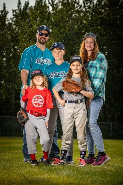 A family of five stands on a baseball field outdoors, with trees in the background. They are dressed in baseball uniforms and casual clothing, with some wearing caps and sunglasses. The children are wearing baseball uniforms and holding a baseball glove, and the adults are standing behind them, smiling.