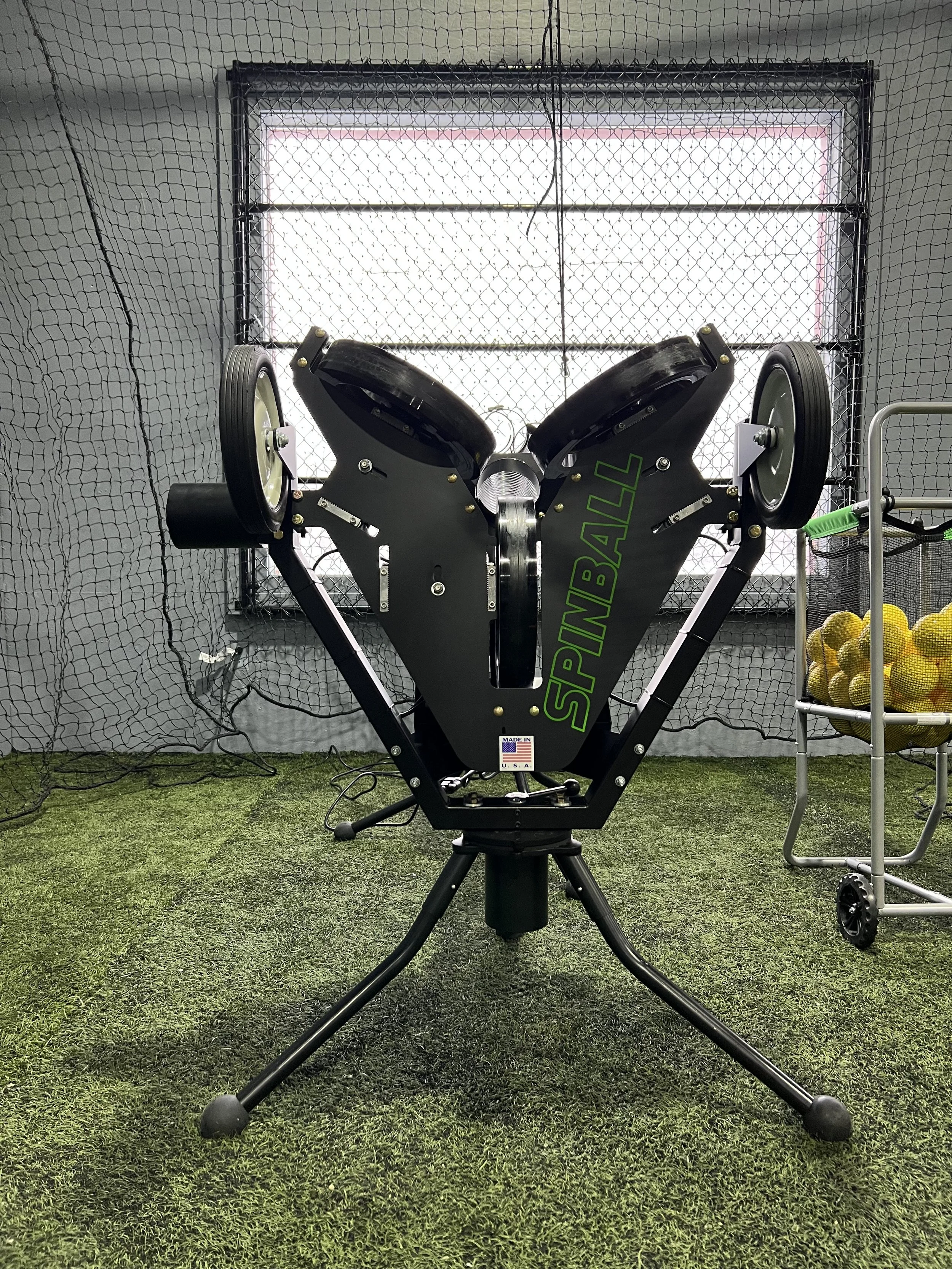 A black Spinballer golf ball return machine with two rotors on top, situated on a green indoor turf with a netted wall and a window in the background, and a cart with yellow tennis balls on the right.