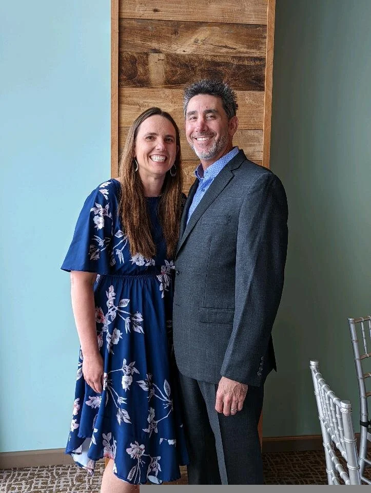 A smiling couple stands together indoors, with a wooden wall behind them and a light blue wall to the left.