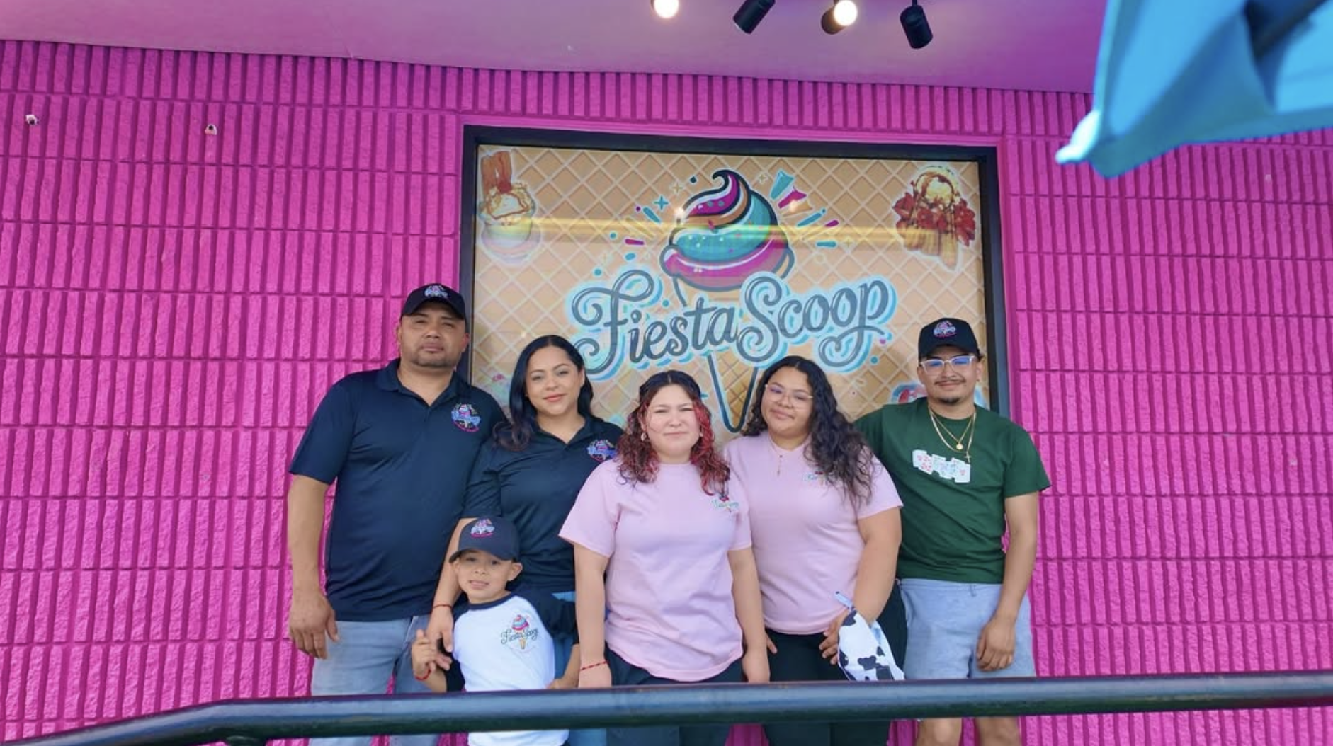 A group of five people, including a young boy, posing in front of a pink wall with a sign reading 'Fiesta Scoop' with a colorful ice cream cone graphic above it.