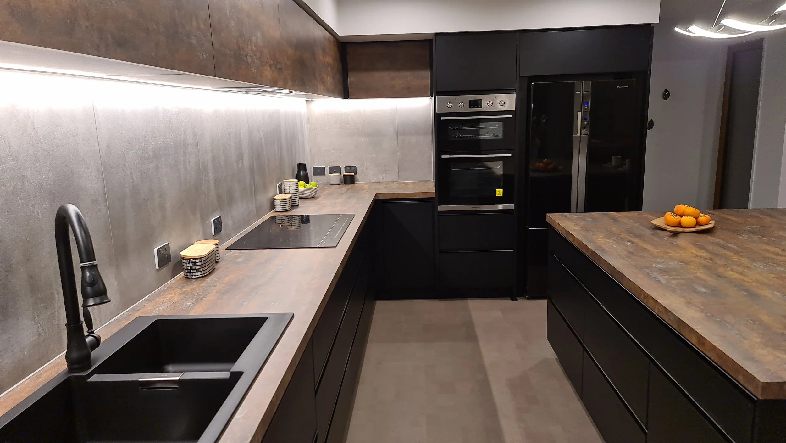 Modern kitchen with black cabinets, wooden countertops, and a concrete backsplash. Contains a black sink, an induction stove, and built-in ovens. In the corner, there are small bowls, a black bottle, and a plate of green apples. On the island, there'