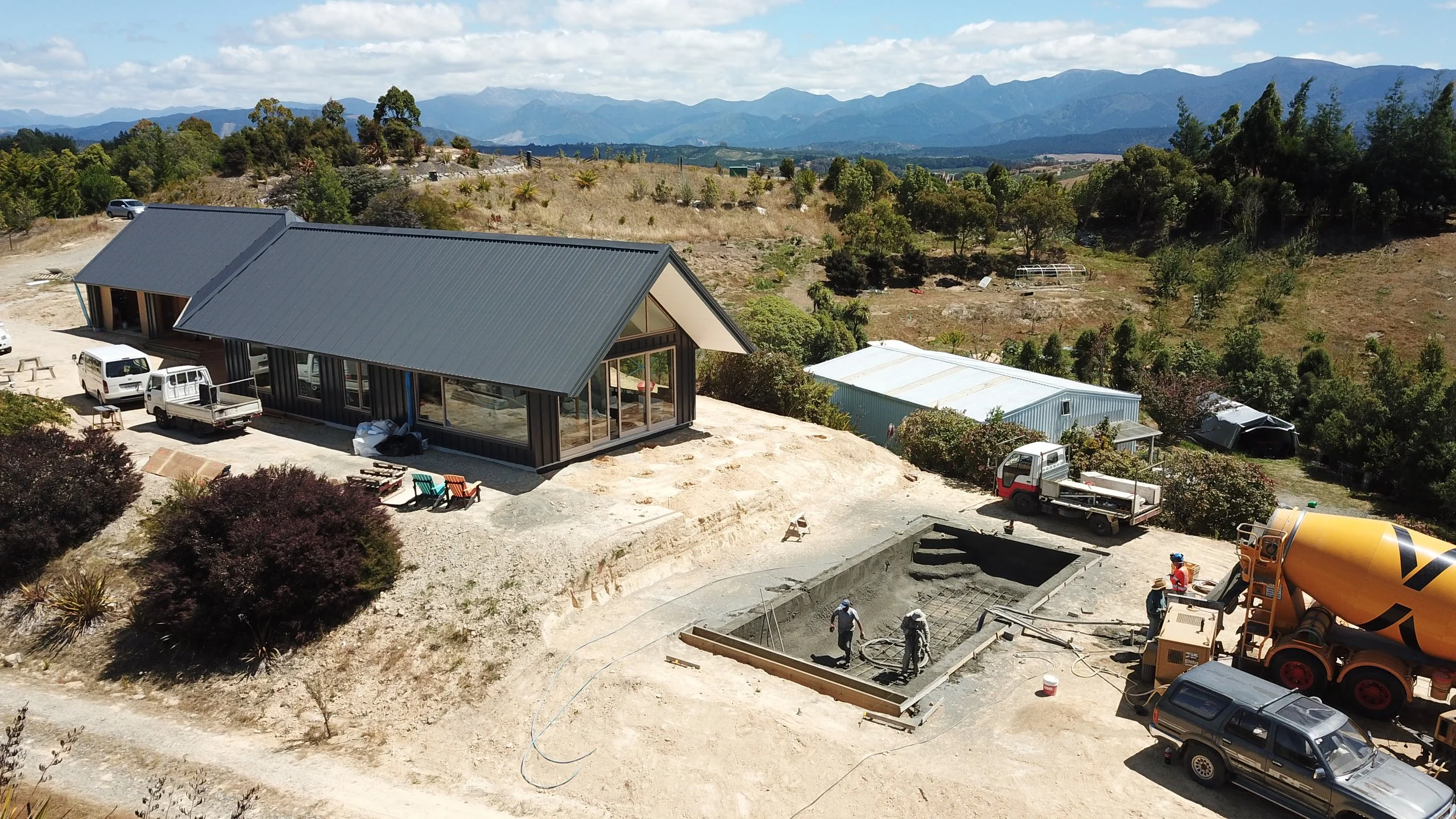 Construction site of a house on a hill with surrounding trucks, construction workers, and a concrete mixer, overlooking mountainous terrain in the background.