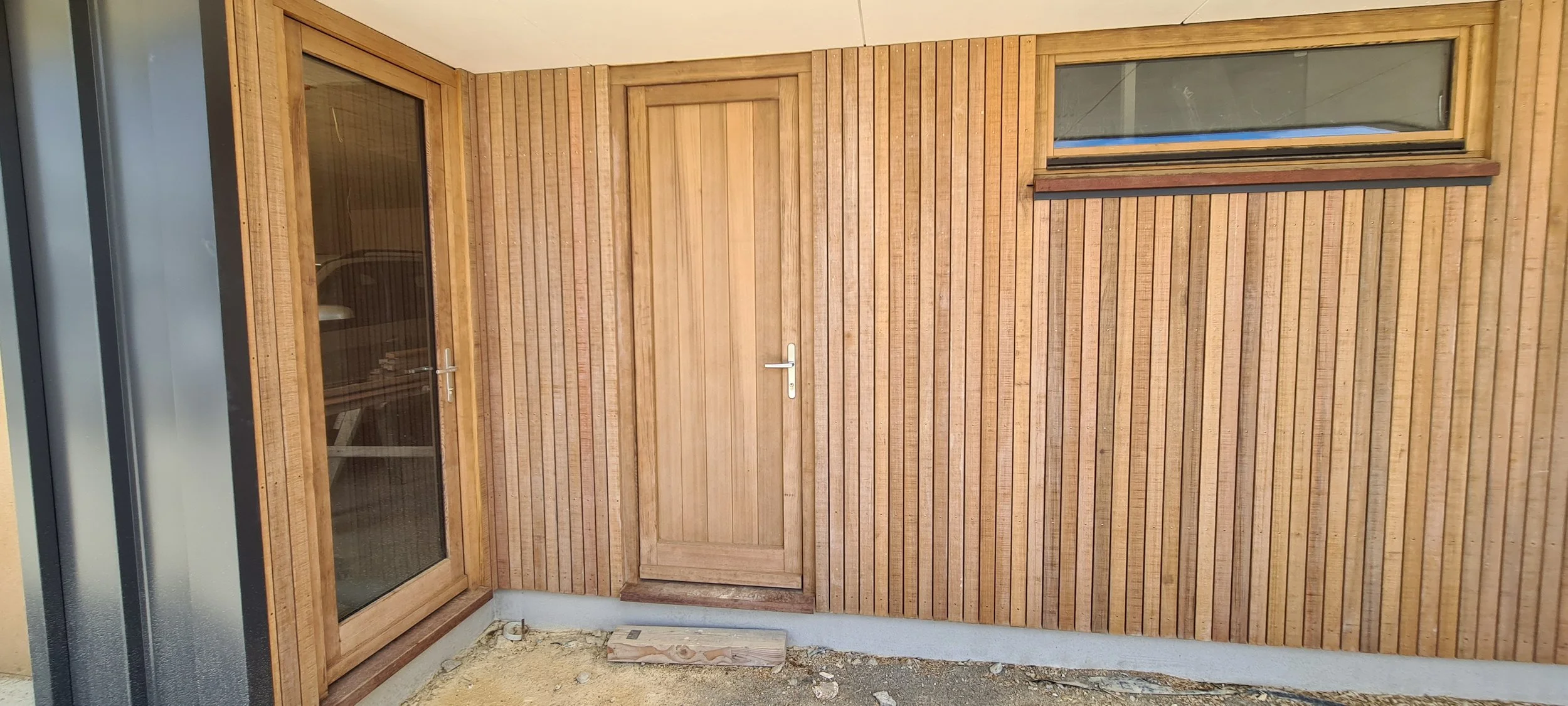 A wooden exterior wall with vertical planks, featuring a glass door with a wooden frame and a second plain wooden door, with a small rectangular window above. Timber decking and cladding