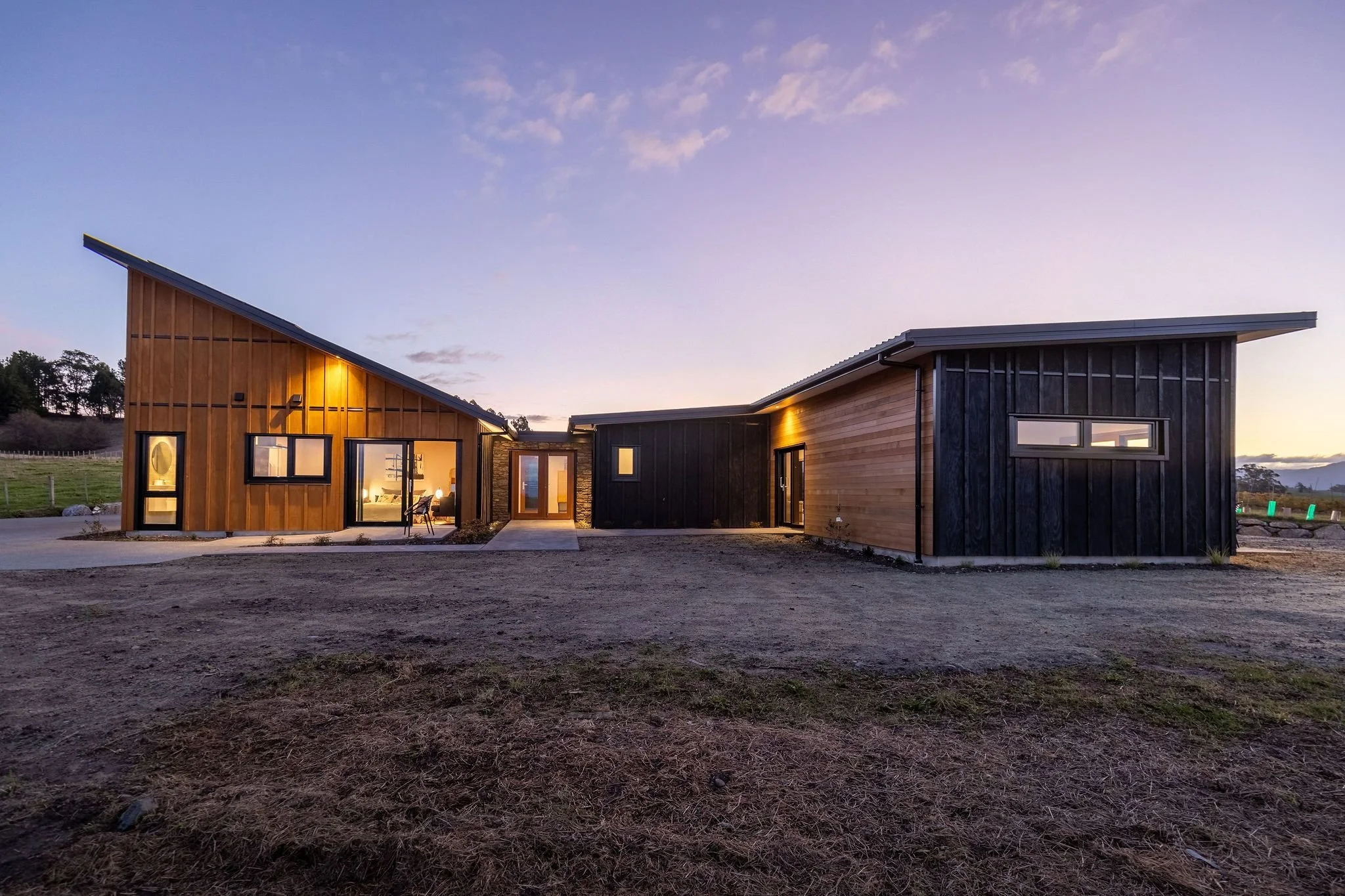 Modern house with a sloped roof, made of wood with black and natural wood panels, illuminated from inside, set against a sunset sky.