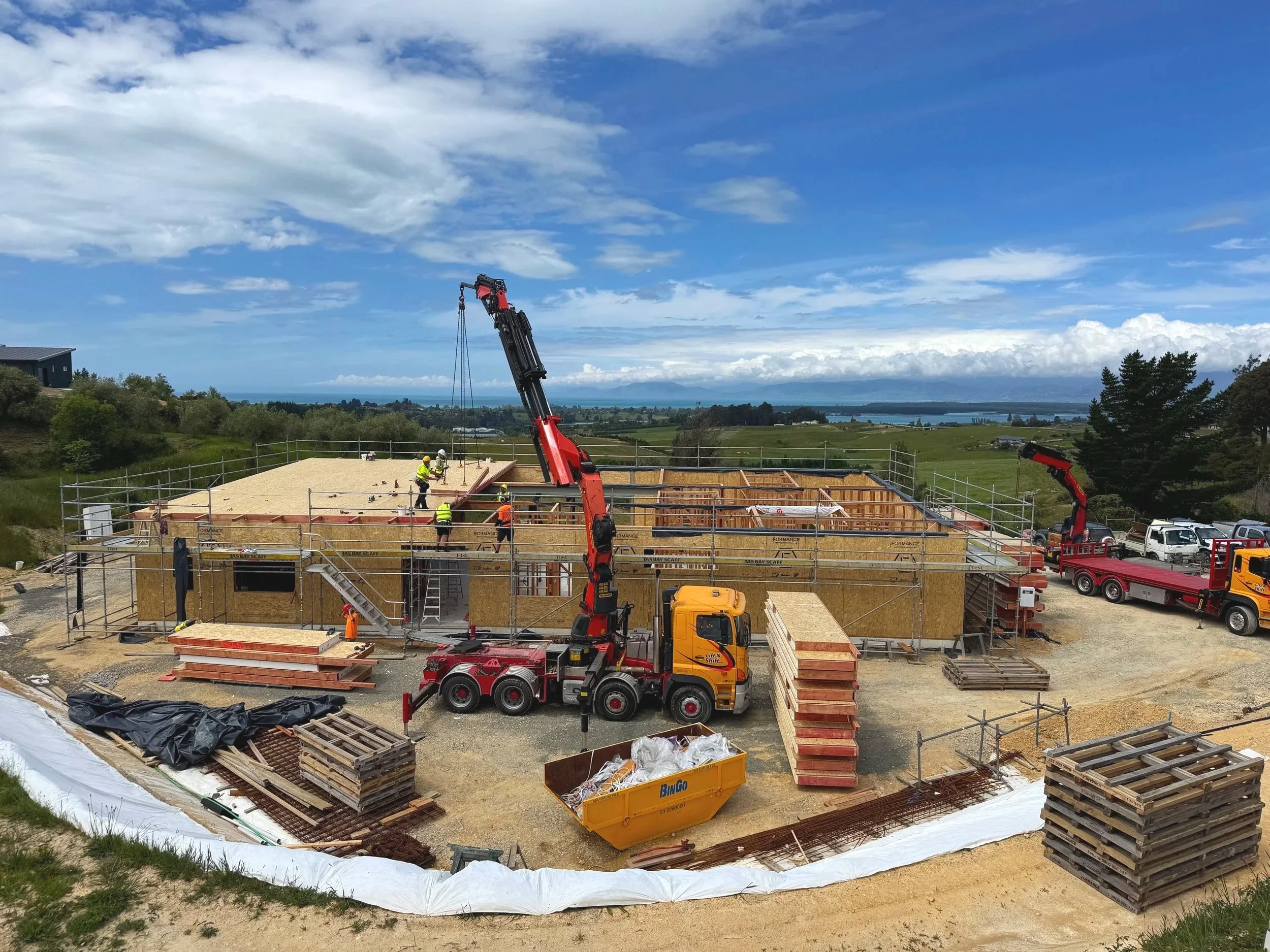 Construction site with workers and cranes building a house on a hill overlooking a landscape with water, trees, and sky.