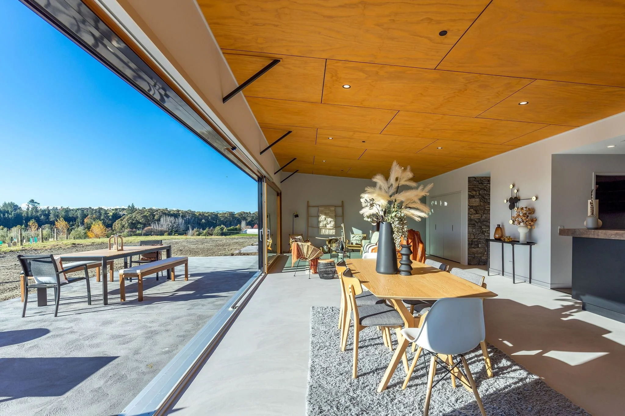 Modern dining area with a large sliding glass door opening to an outdoor patio with lawn and trees, featuring a wooden dining table with nine chairs, a vase with pampas grass, and a living space with furniture and decor.