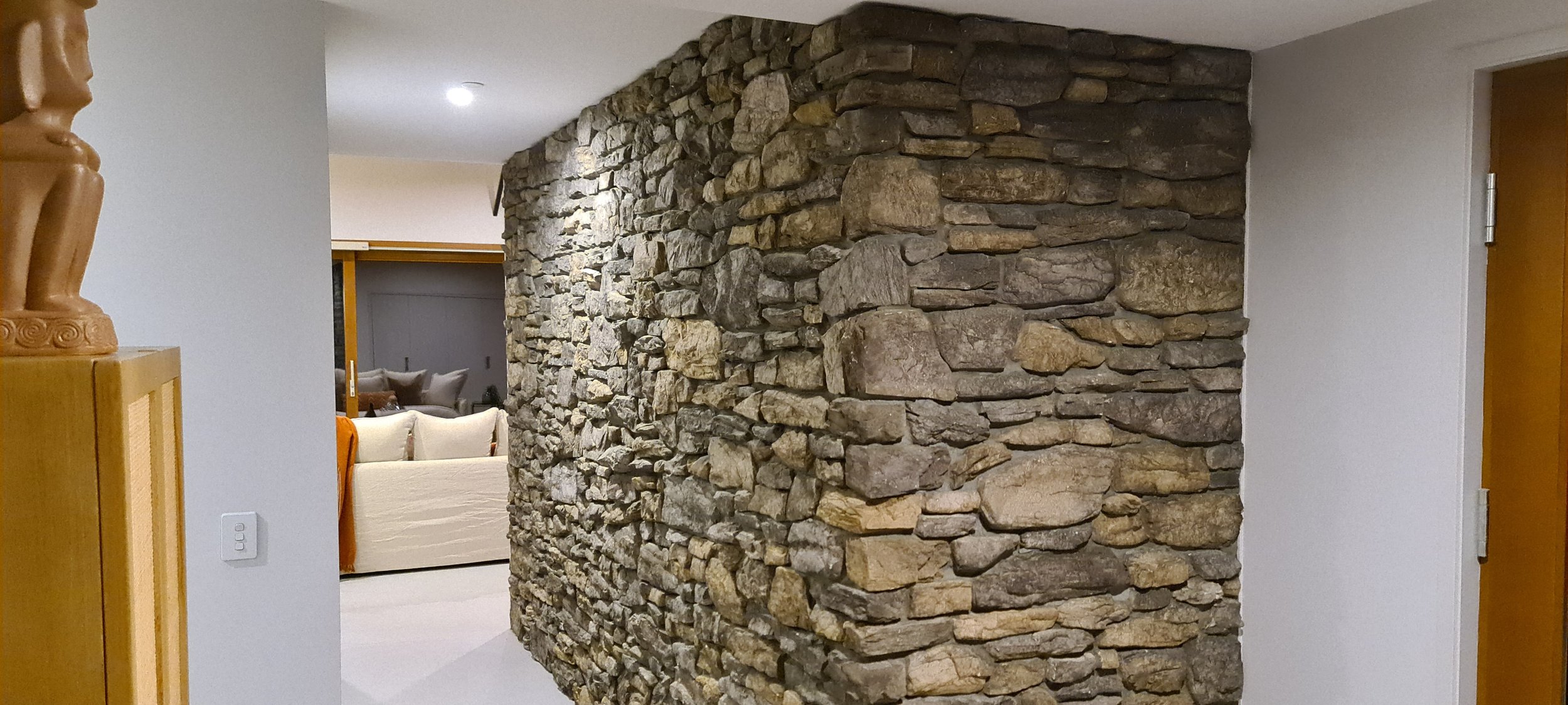 Interior view of a home showing a curved stone wall dividing the space, with white walls and a doorway on the right. In the background, a living room with a beige sofa and cushions is visible.