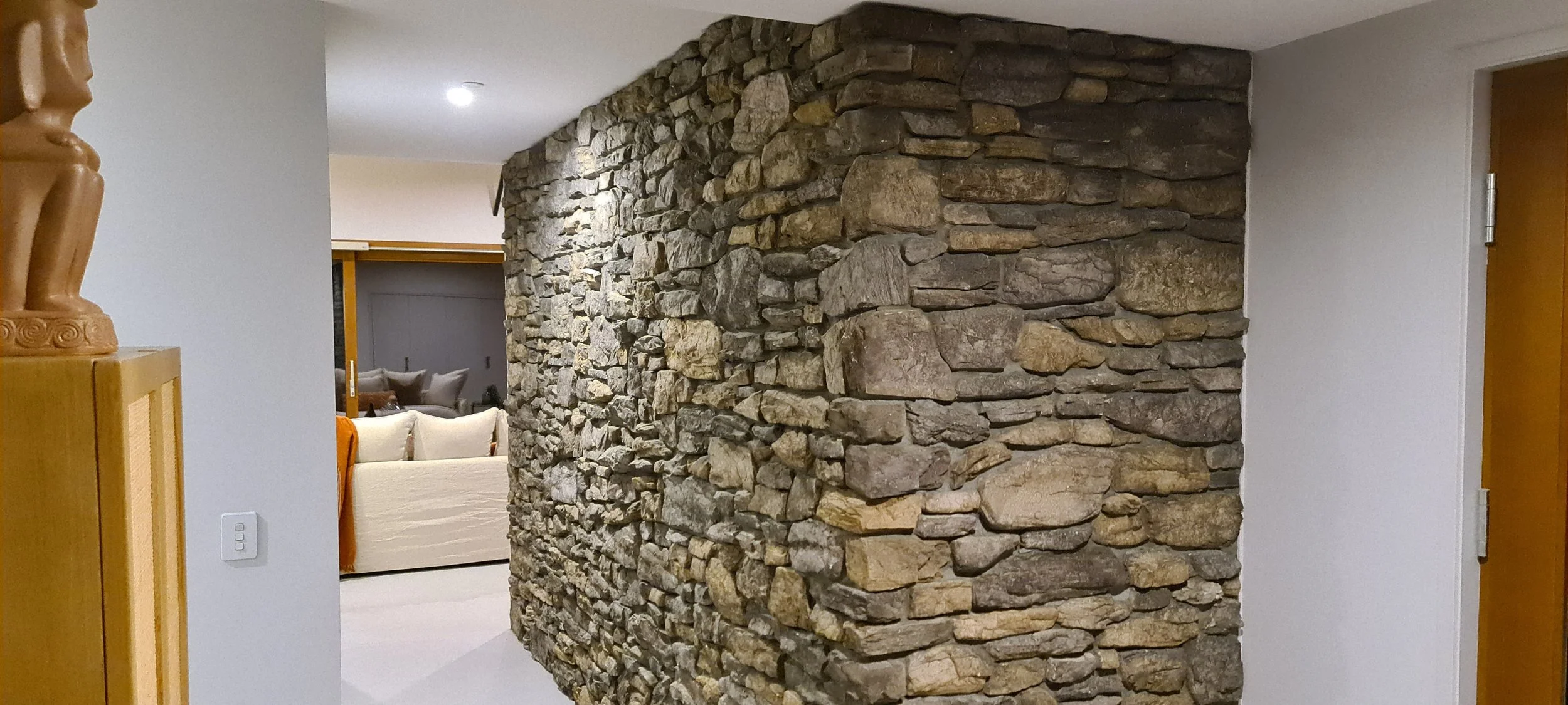 Interior view of a home showing a curved stone wall dividing the space, with white walls and a doorway on the right. In the background, a living room with a beige sofa and cushions is visible.