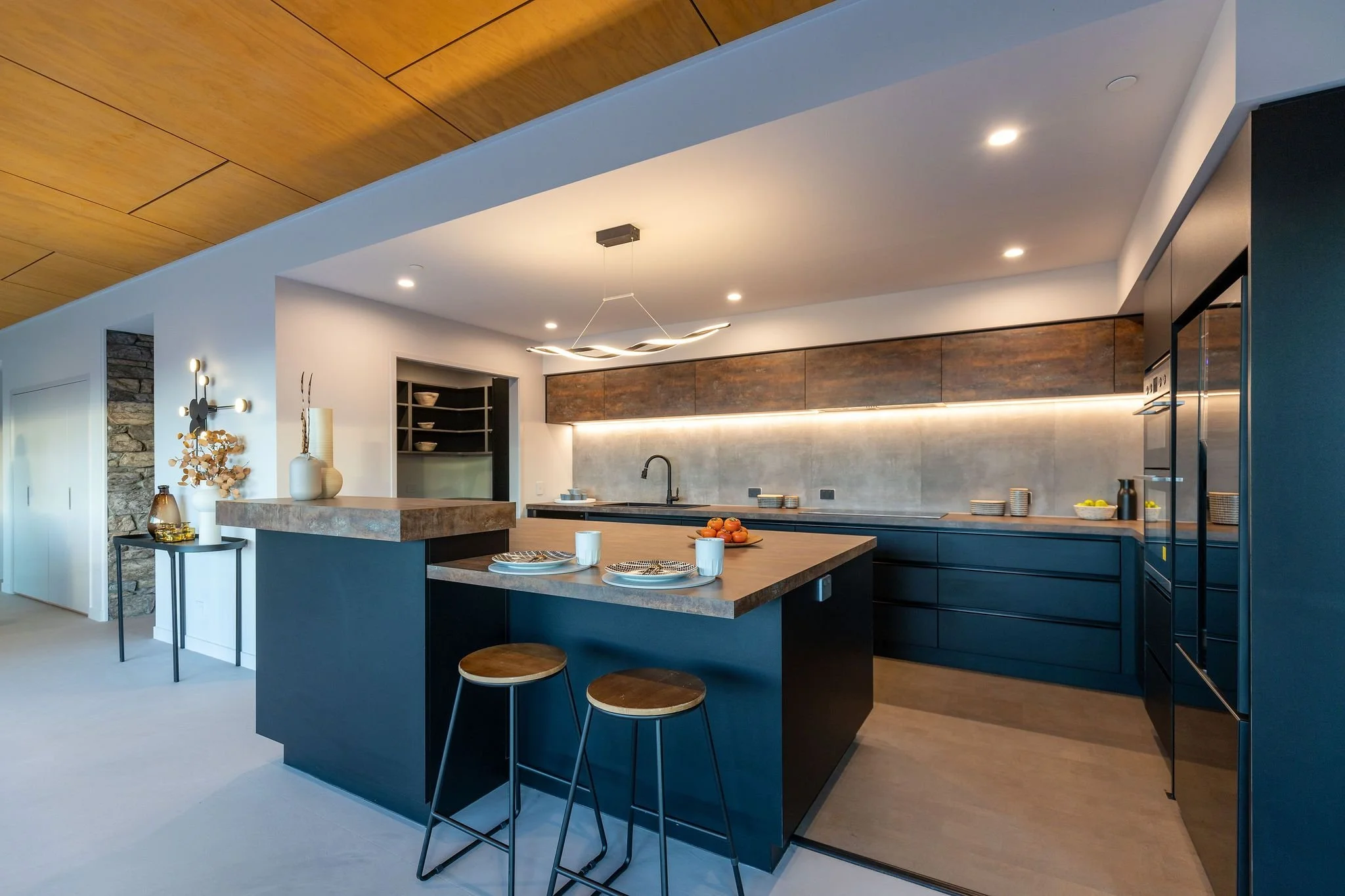Modern kitchen with navy blue cabinets, a large island with a wooden countertop, two stools, and contemporary lighting; a bowl of oranges on the island, minimalist decor, and sleek design elements.