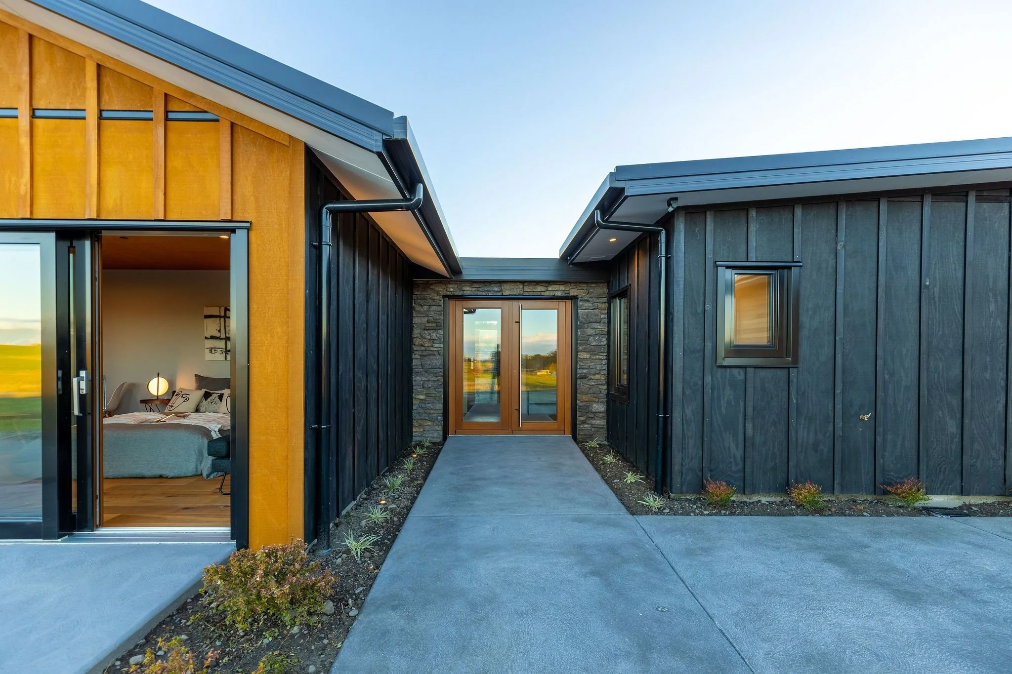 Exterior view of a modern house with black wooden siding and a stone entrance, featuring a glass door with wooden trim. Inside, a bedroom is visible through the left sliding glass door, with a bed, nightstand, and lamp.