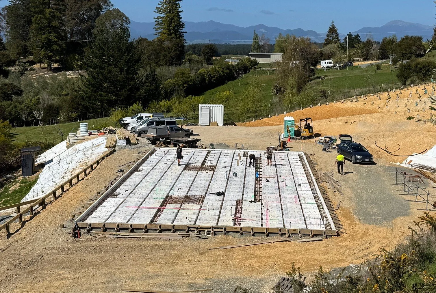 Construction site showing a foundation for a building with workers, construction vehicles, and parked cars around, with trees and mountains in the background.