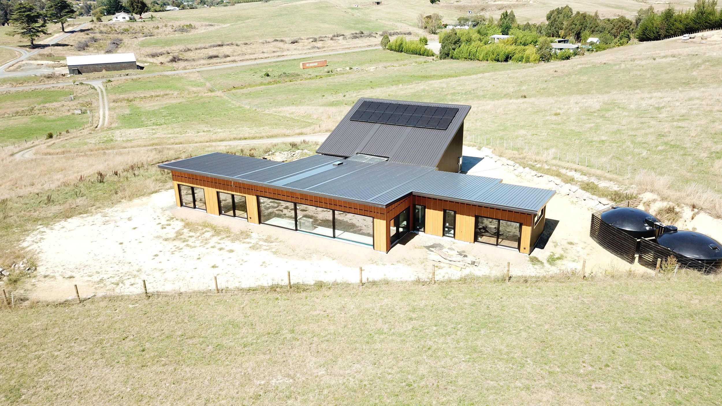 A modern house with a metal roof, large windows, and outdoor solar panels in a rural landscape with grassy fields and a few scattered trees.