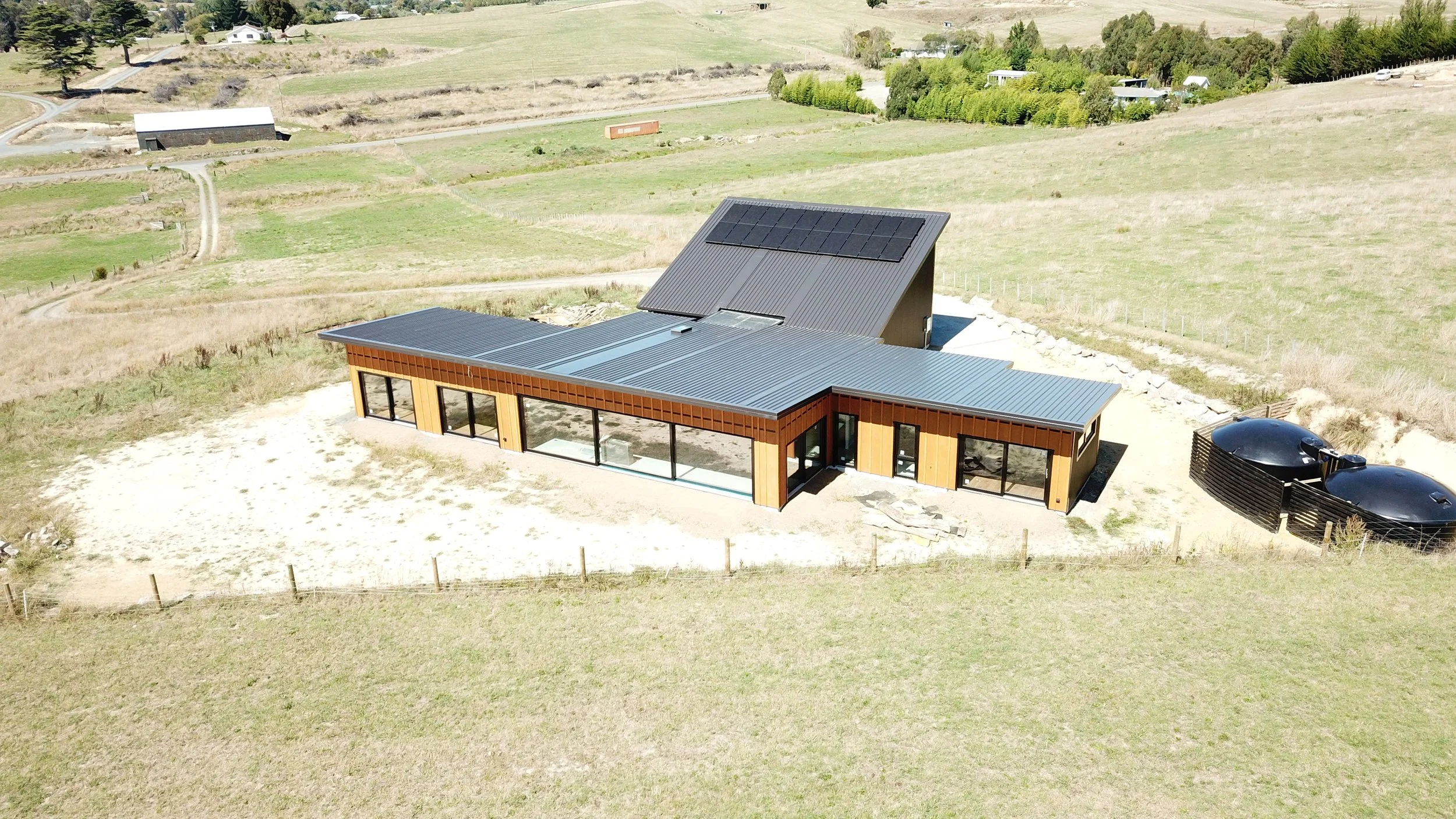 A modern house with a metal roof, large windows, and outdoor solar panels in a rural landscape with grassy fields and a few scattered trees.