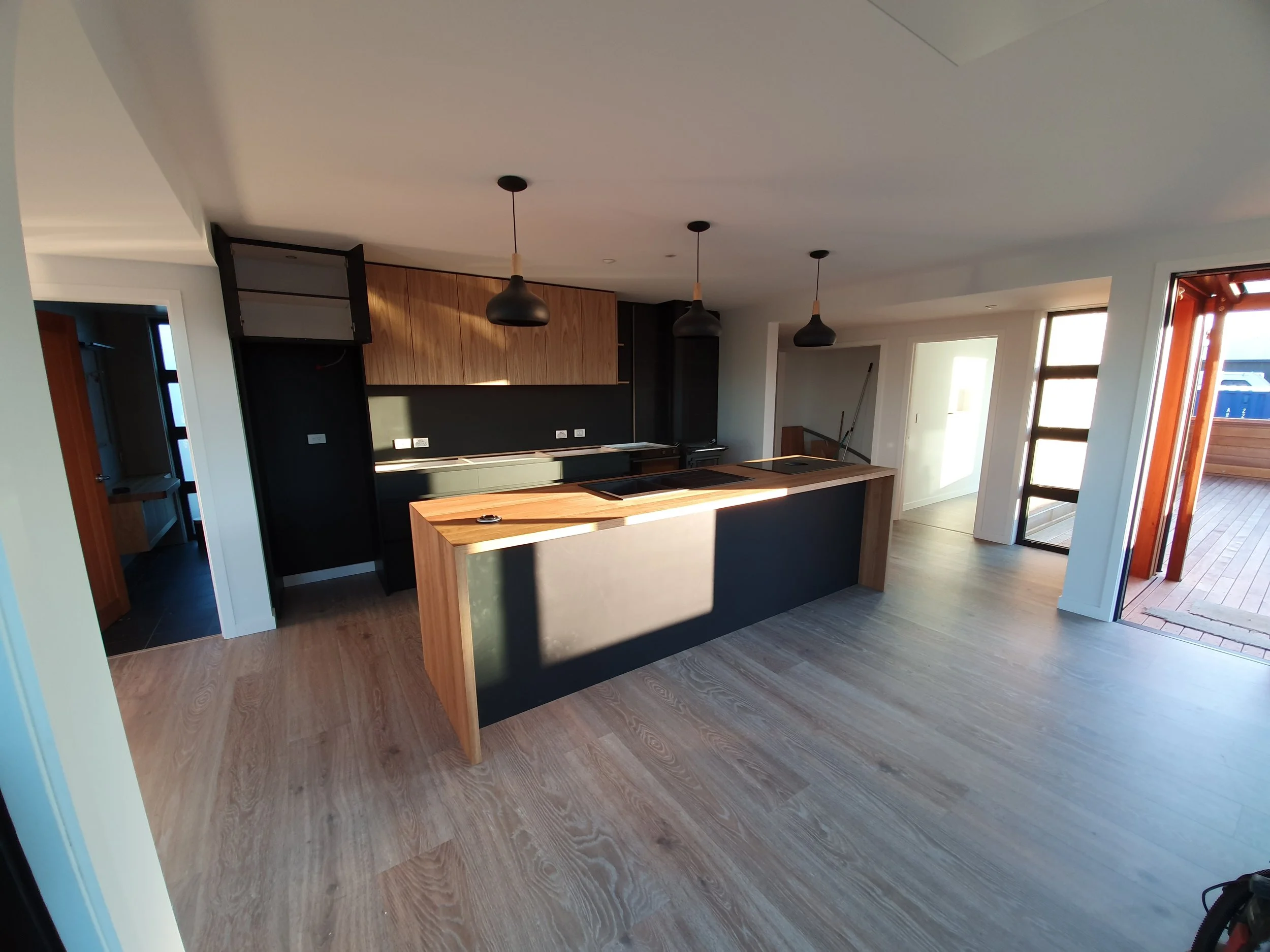 Modern kitchen with black and wooden cabinetry, a large wooden island, pendant lights, and sliding glass doors leading to an outdoor deck.