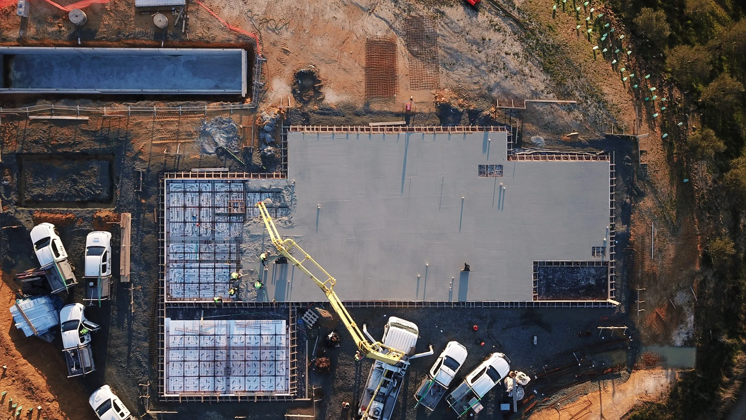 Aerial view of a construction site with workers pouring concrete for the foundation of a building. Several trucks and construction vehicles are present, and the framework of the building is partially completed.