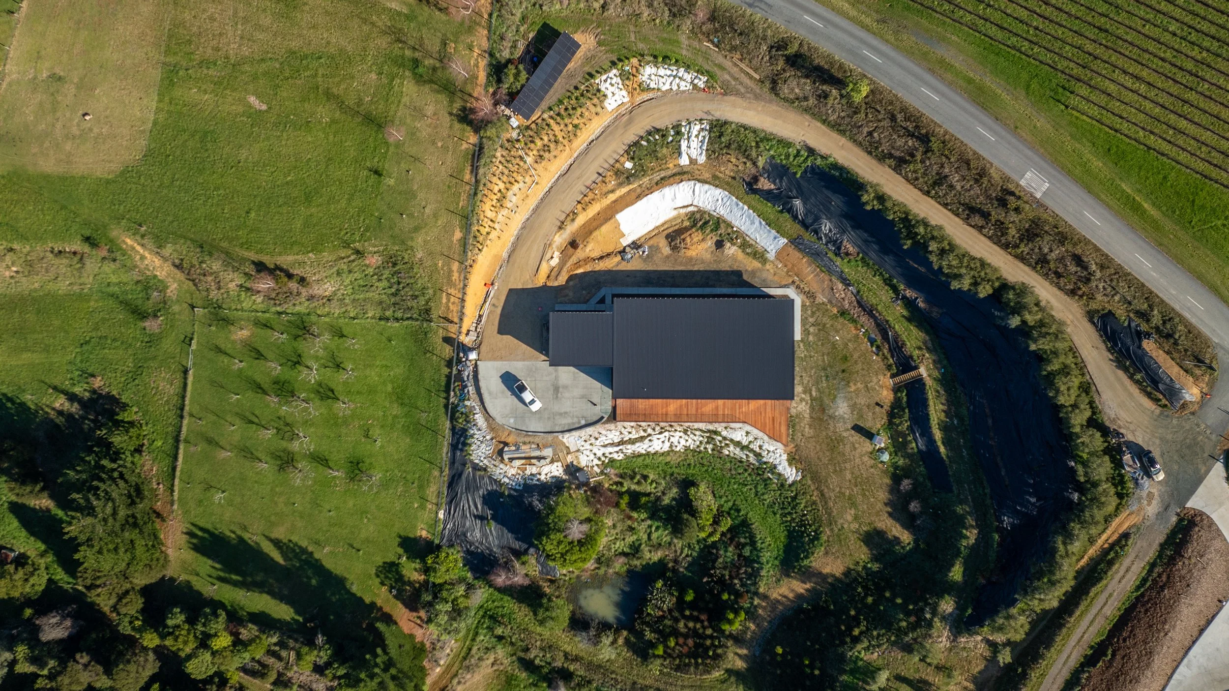 Aerial view of a house under construction with a black roof, surrounded by green fields, trees, and a curved dirt driveway, with construction materials and equipment nearby.