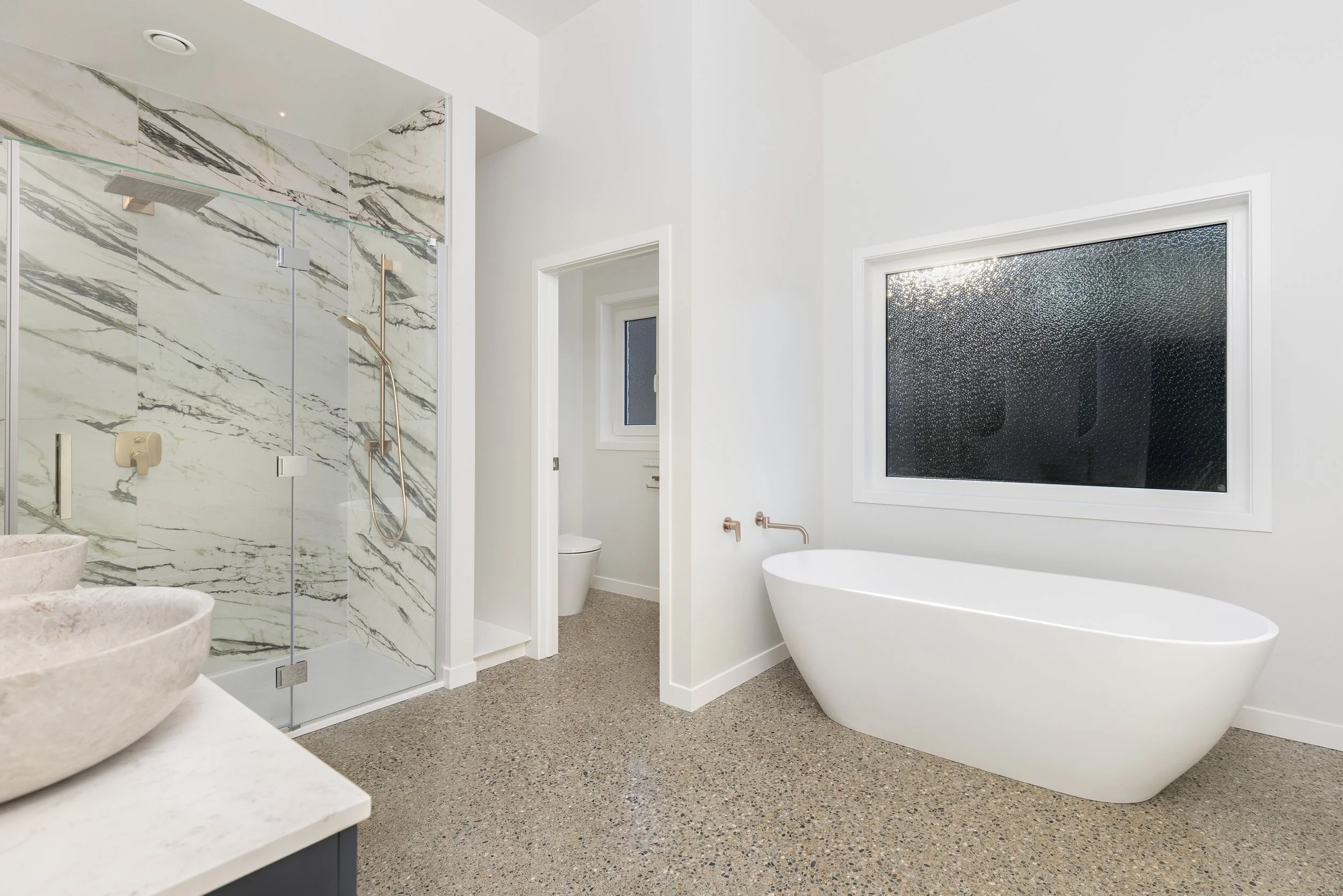 Modern bathroom with a freestanding bathtub, a large frosted window, a shower with marble walls, and a separate toilet room.