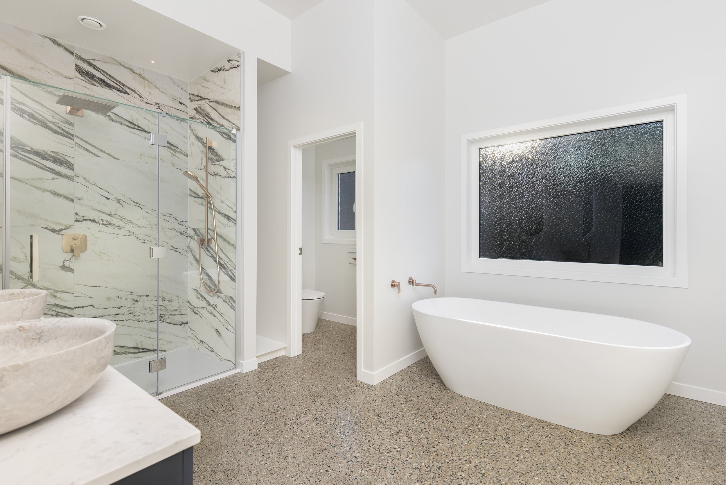 Modern bathroom with a freestanding bathtub, a large frosted window, a shower with marble walls, and a separate toilet room.