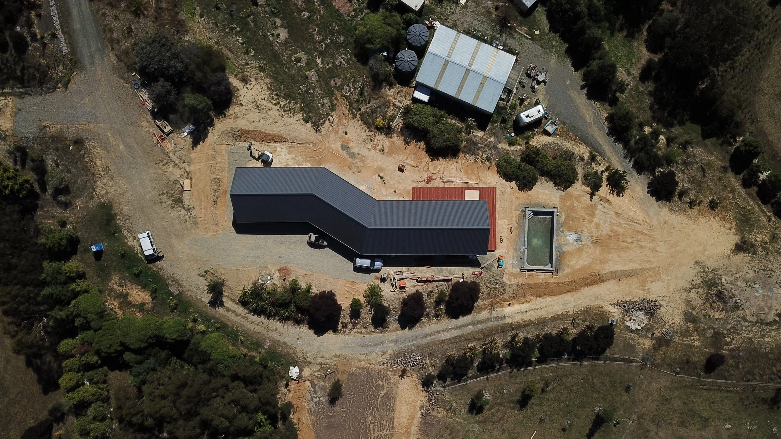 An aerial view of a construction site with a dark gray building, a swimming pool, a gravel driveway, and surrounding trees and vegetation.