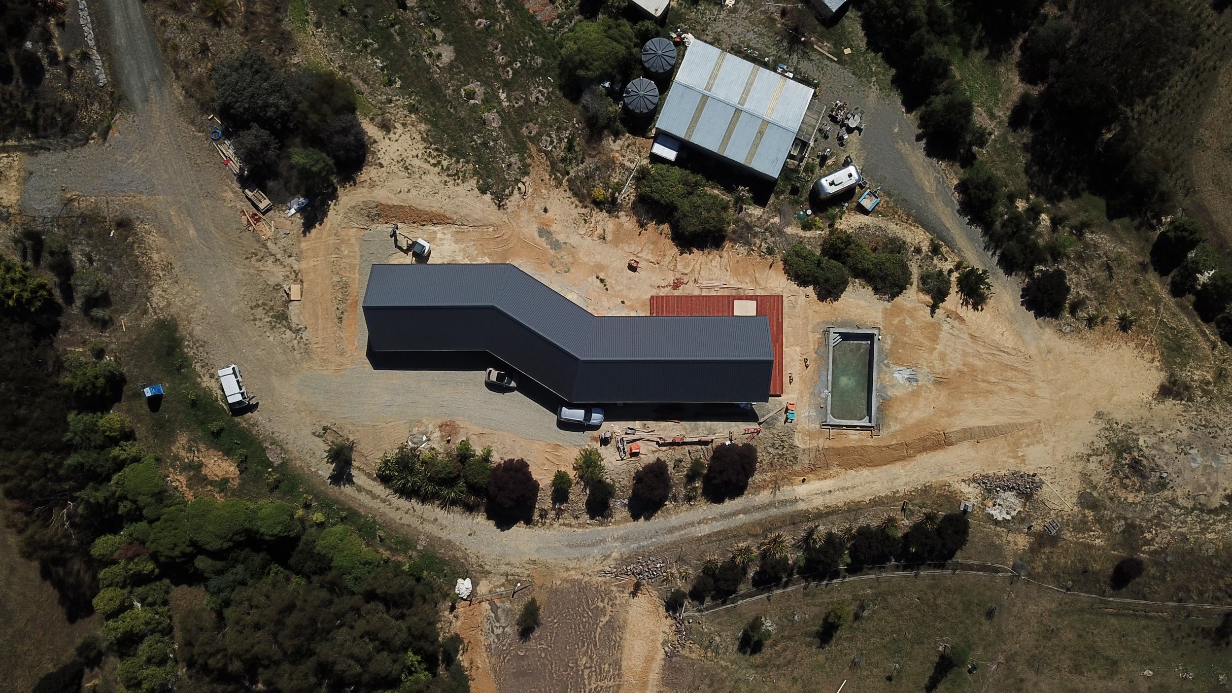 An aerial view of a construction site with a dark gray building, a swimming pool, a gravel driveway, and surrounding trees and vegetation.