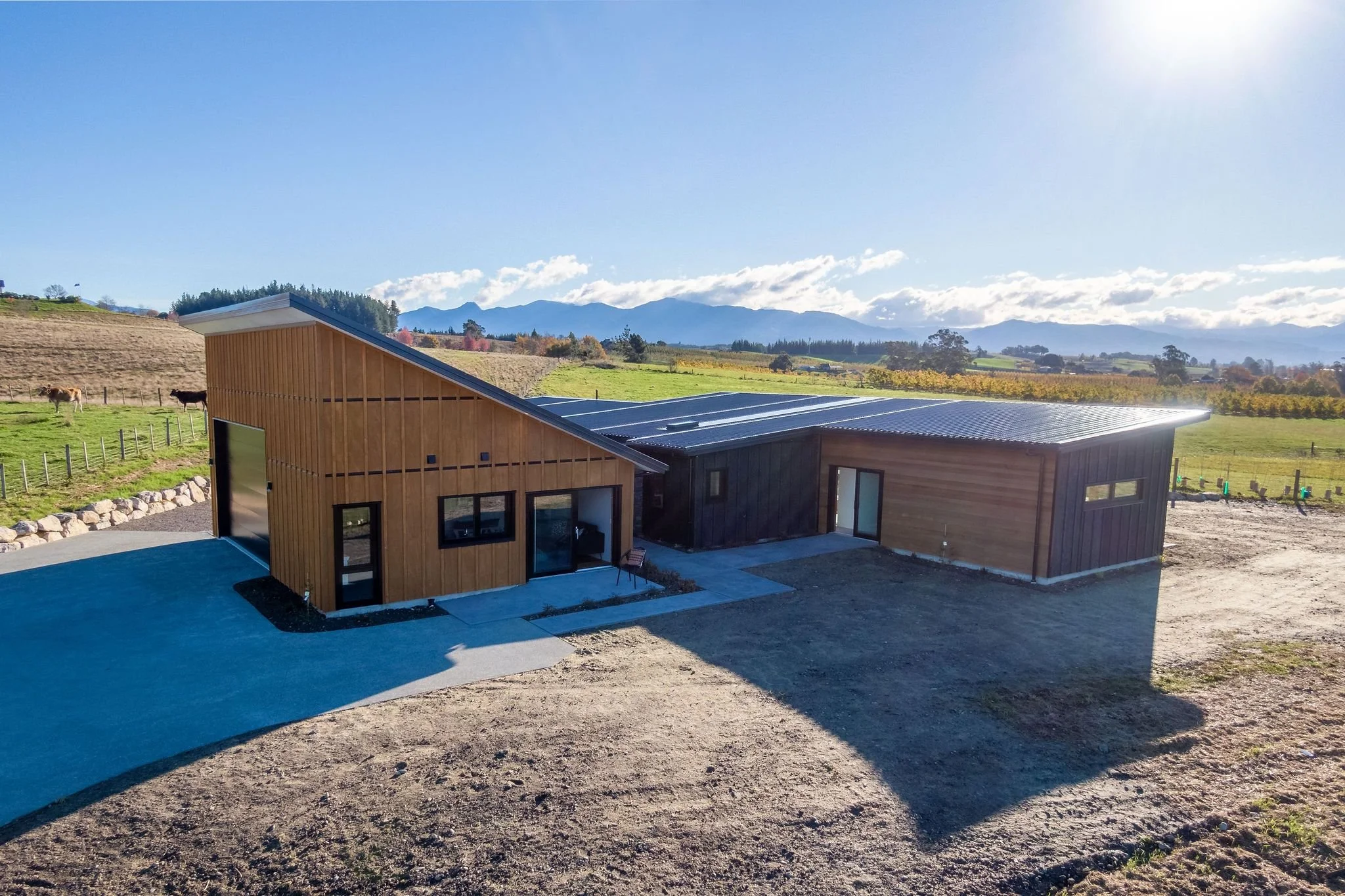 Modern barn-style building with wooden exterior, large metal roof, and glass doors, set on a rural landscape with fields, mountains, and blue sky.