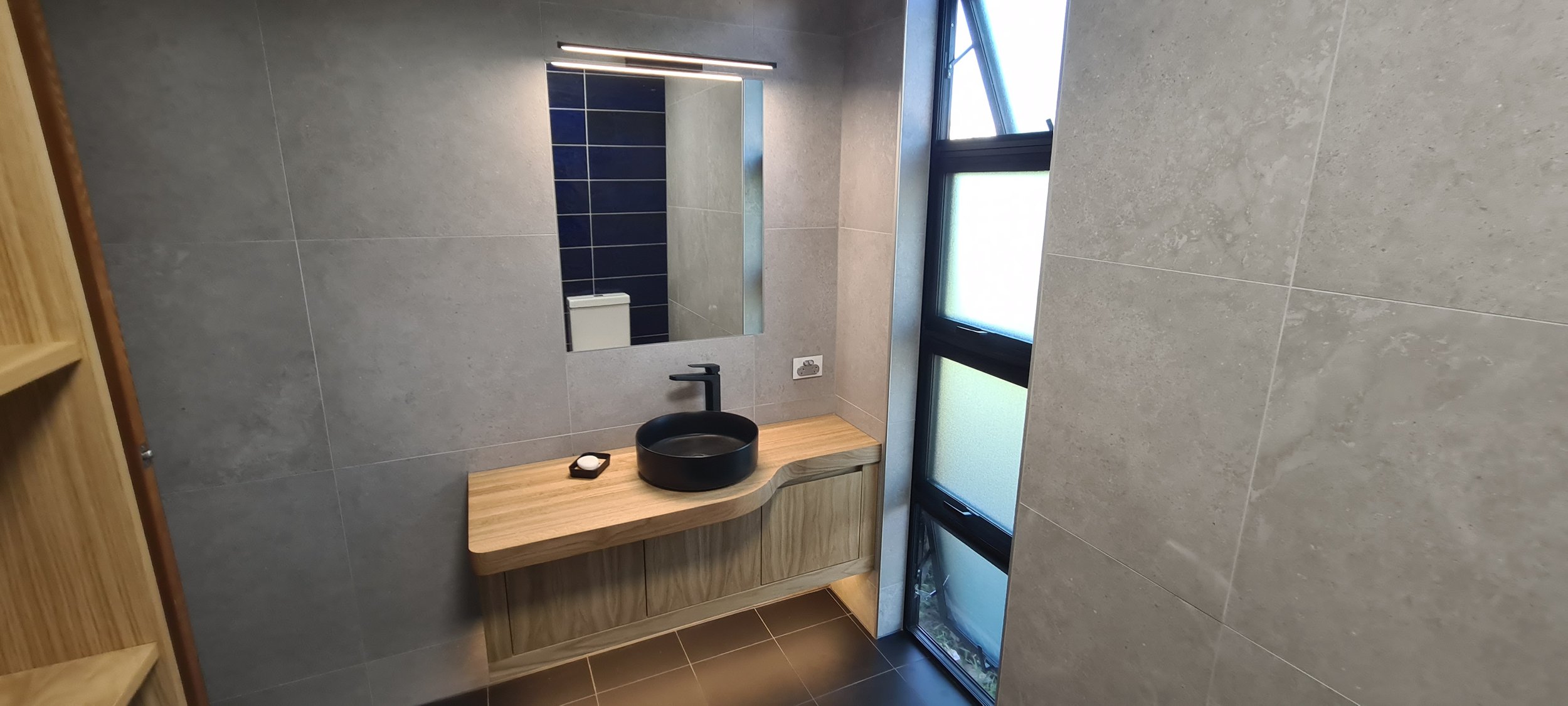 Modern bathroom with a round black sink on a wooden vanity, a mirror above, and frosted glass window panels letting in natural light.