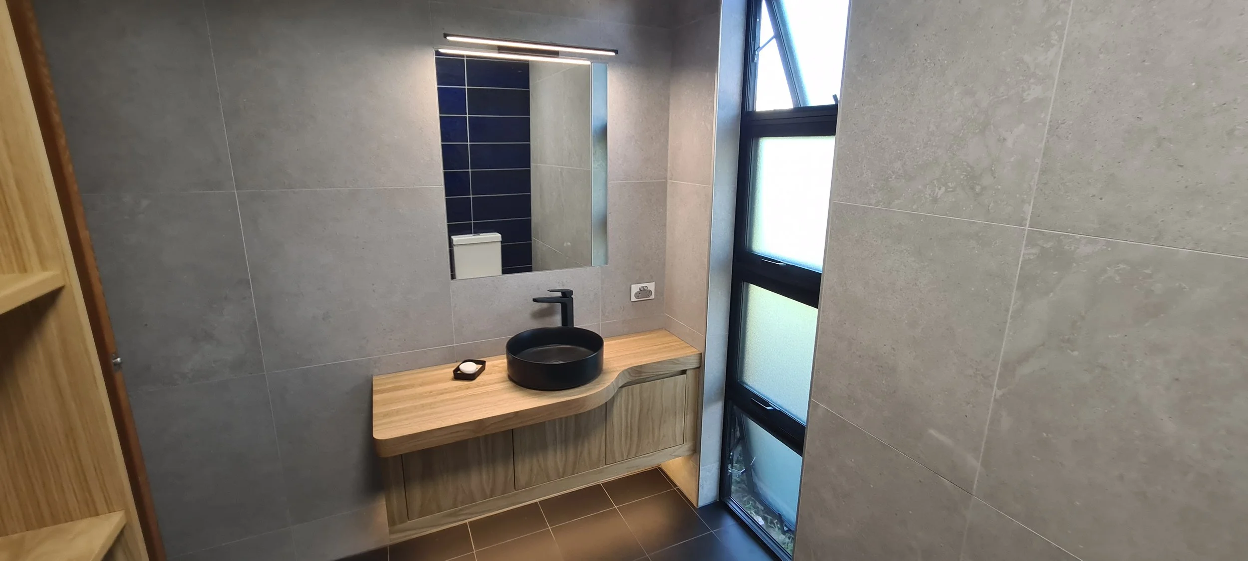 Modern bathroom with a round black sink on a wooden vanity, a mirror above, and frosted glass window panels letting in natural light.